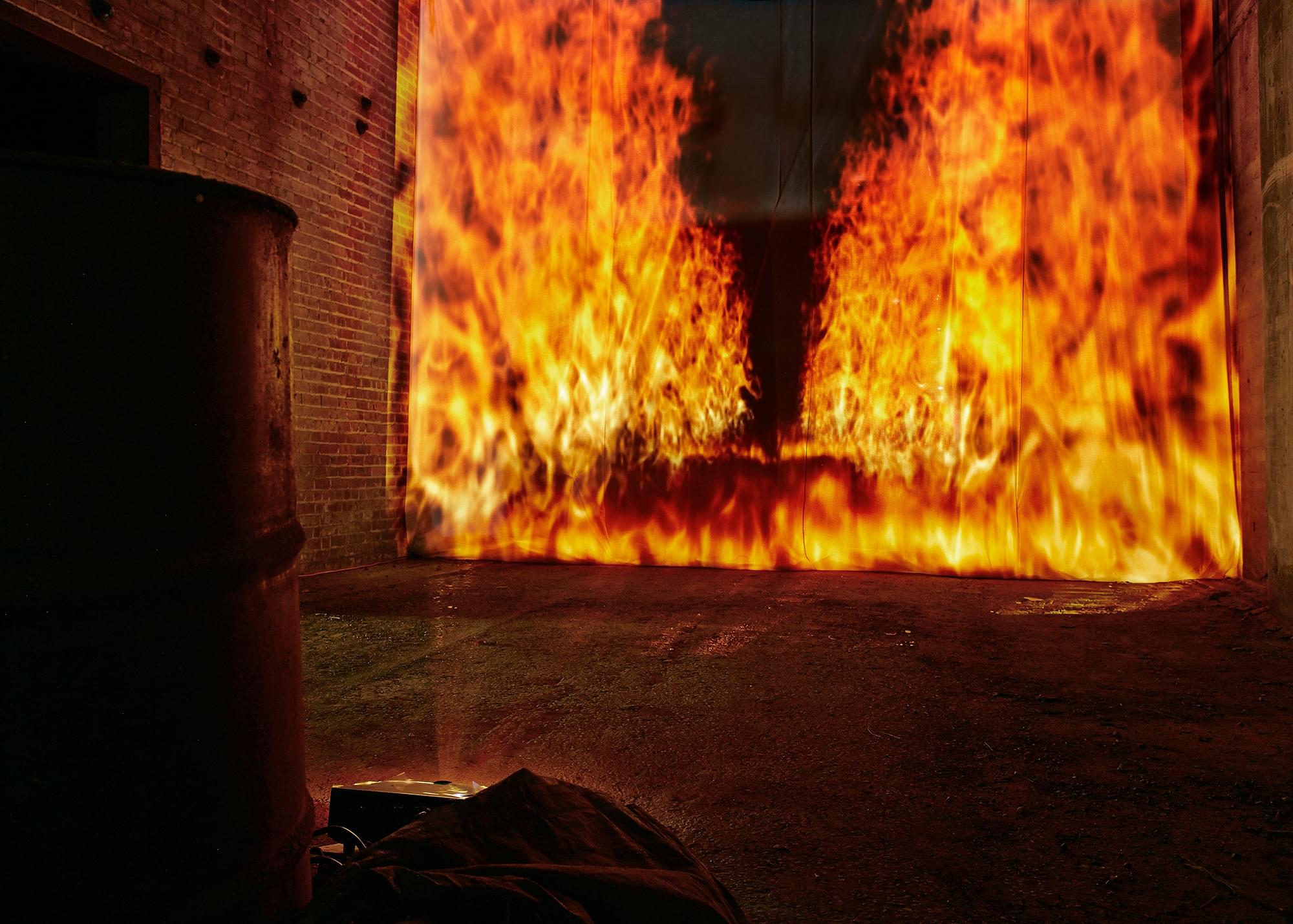 Close-up nighttime photograph of an oil drum on the left and a (projected) raging firescape in the background towards the right