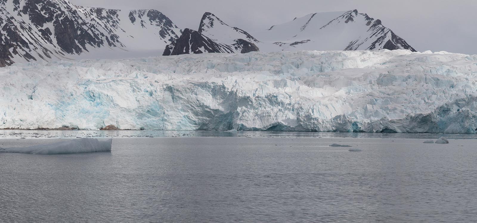 Panoramic photograph two of five: the ice island has shifted to be almost out of frame on the left. Glacier in the background and snow covered mountains behind it.