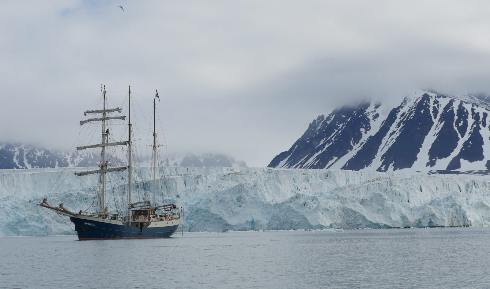 Photograph of an arctic landscape: cold gray sky, snow-covered mountains in the far background, a 20-30 meter high glacier in the near-background, and a old-timey sail ship anchored in calm waters on the left of the frame.