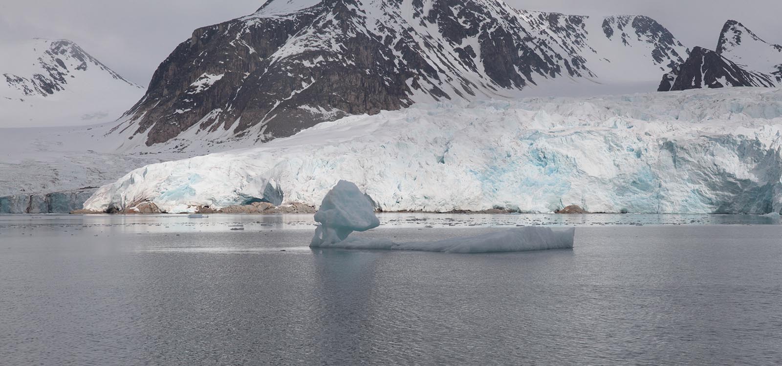 Panoramic photograph one of five that forms the leftmost side of an Arctic landscape. In the foreground is a steel gray body of water. In the middle of the frame is a tiny ice-island that looks like it probably calved off a glacier. In the background is a 20-meter-ish high glacier with snow-capped mountains behind it.