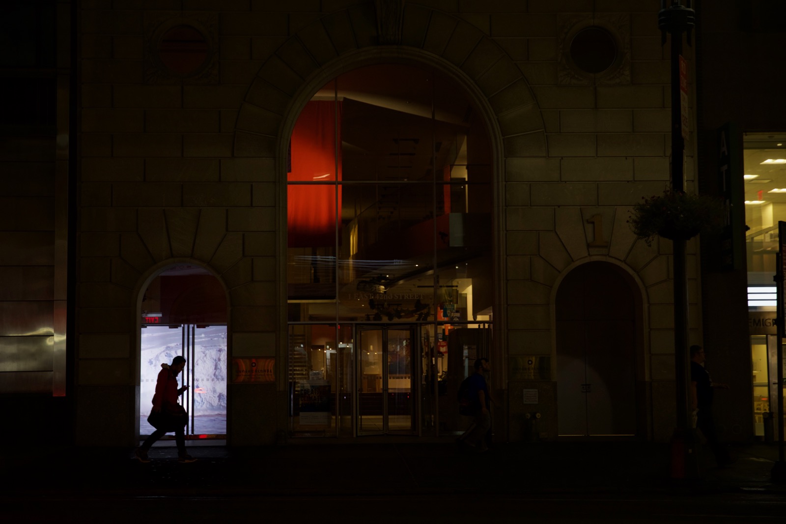 Small thumbnail image of Very dark nighttime photograph of a silhouetted figure walking down the street looking at their phone. Behind him is a projected image of glaciers in a small storefront window.