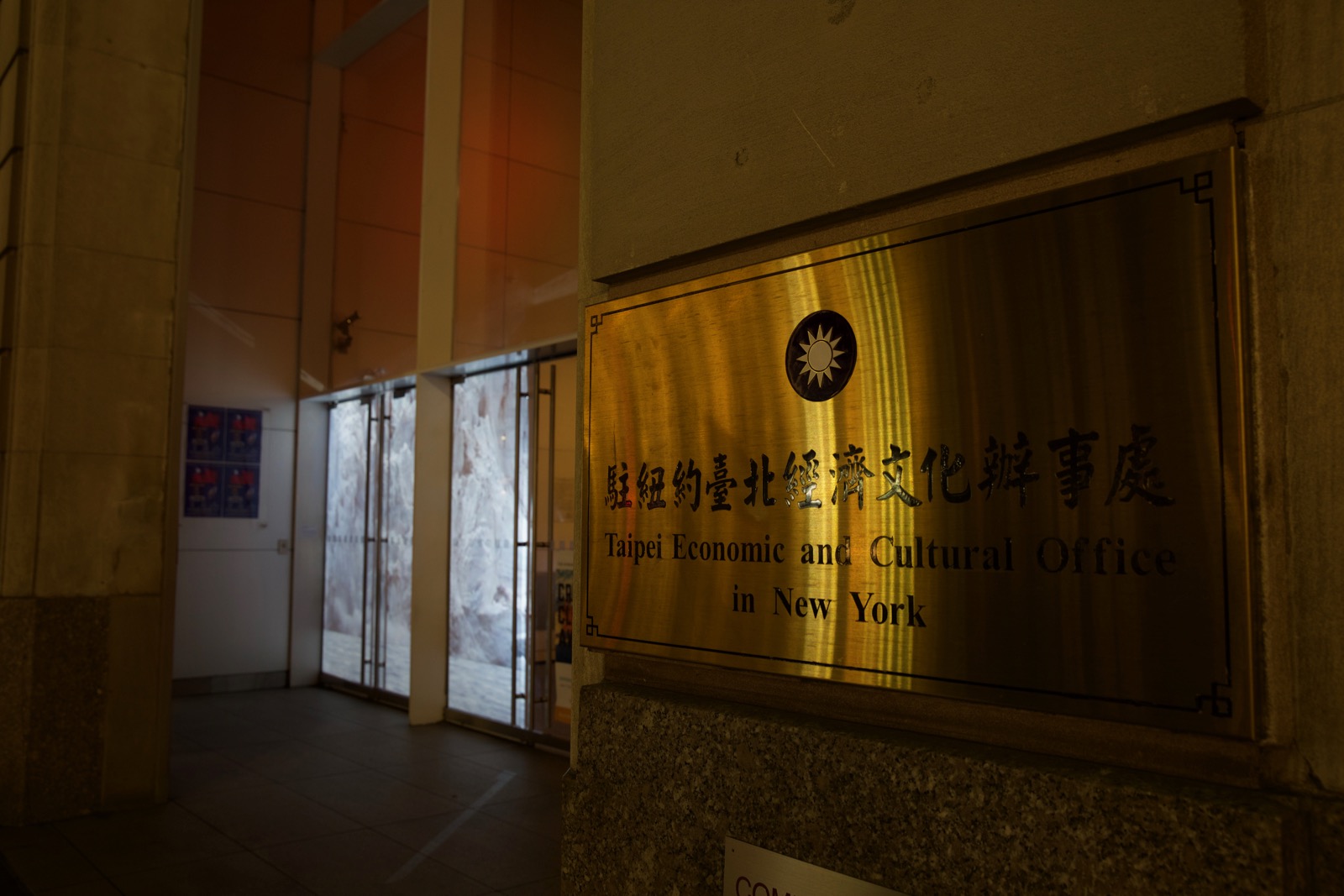 Close up nighttime photograph of the signage of the building in the previous photograph. The signage (a medium-sized gold plate) reads 'Taipei Economic and Cultural Office in New York.'  Blurred in the background is a projection of glaciers in the building's storefront windows.