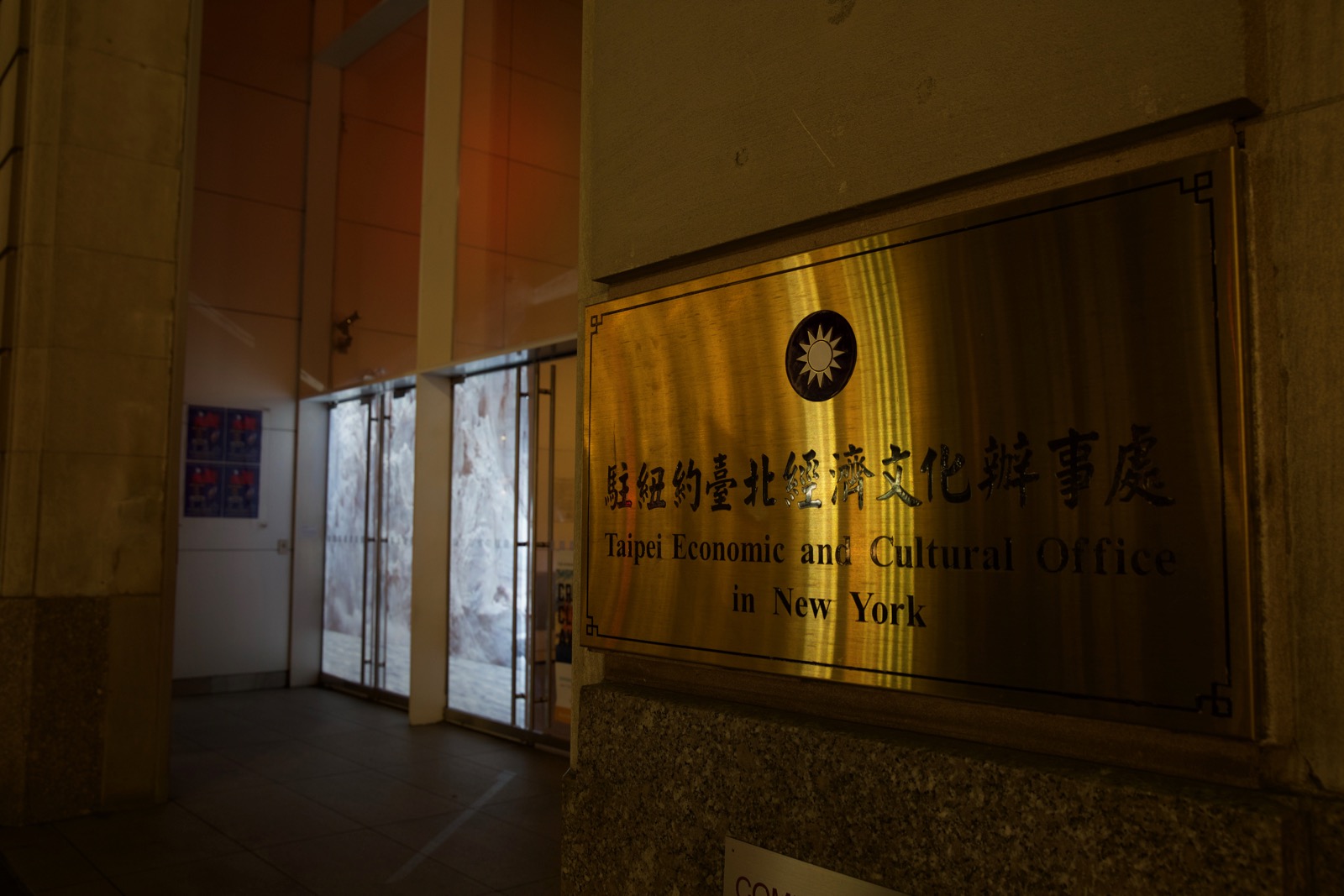 Close up nighttime photograph of the signage of the building in the previous photograph. The signage (a medium-sized gold plate) reads 'Taipei Economic and Cultural Office in New York.' Blurred in the background is a projection of glaciers in the building's storefront windows.