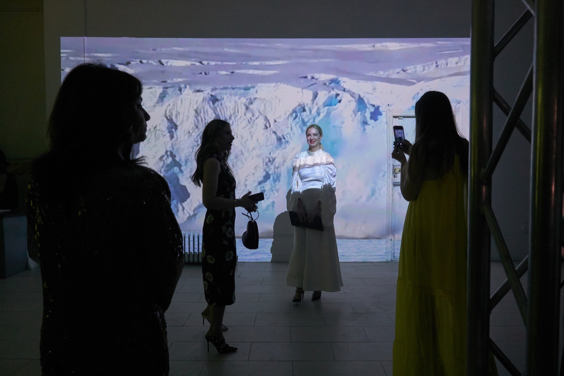 Photograph of four people in a dark gallery space. Only one is illuminated, who appears to be a blond-haired woman, very nicely done-up, and who is having her picture taken by someone else in the frame. She is illuminated by a projection of a glacier collapsing behind her. She is smiling.