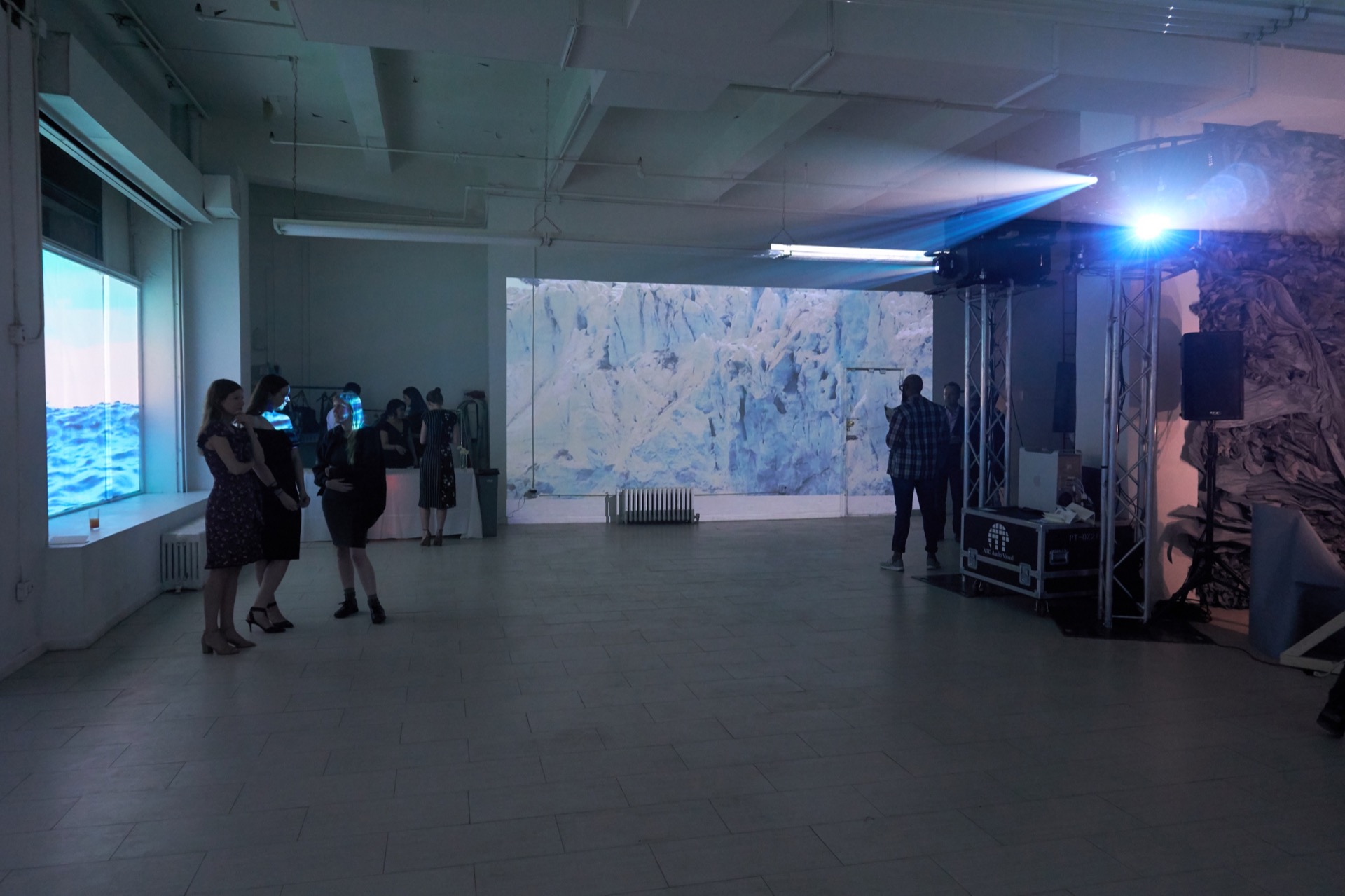 Wide shot of the inside of a gallery showing two different projected works. On the left, an image of a body of water is projected into the gallery windows. Straight back, an image of an Arctic scene&mdash;close up of a glacier&mdash;is projected. There are about a dozen people in cocktail attire milling about. On the right, a two sharply bright lights are emanating from the lens of large projectors.