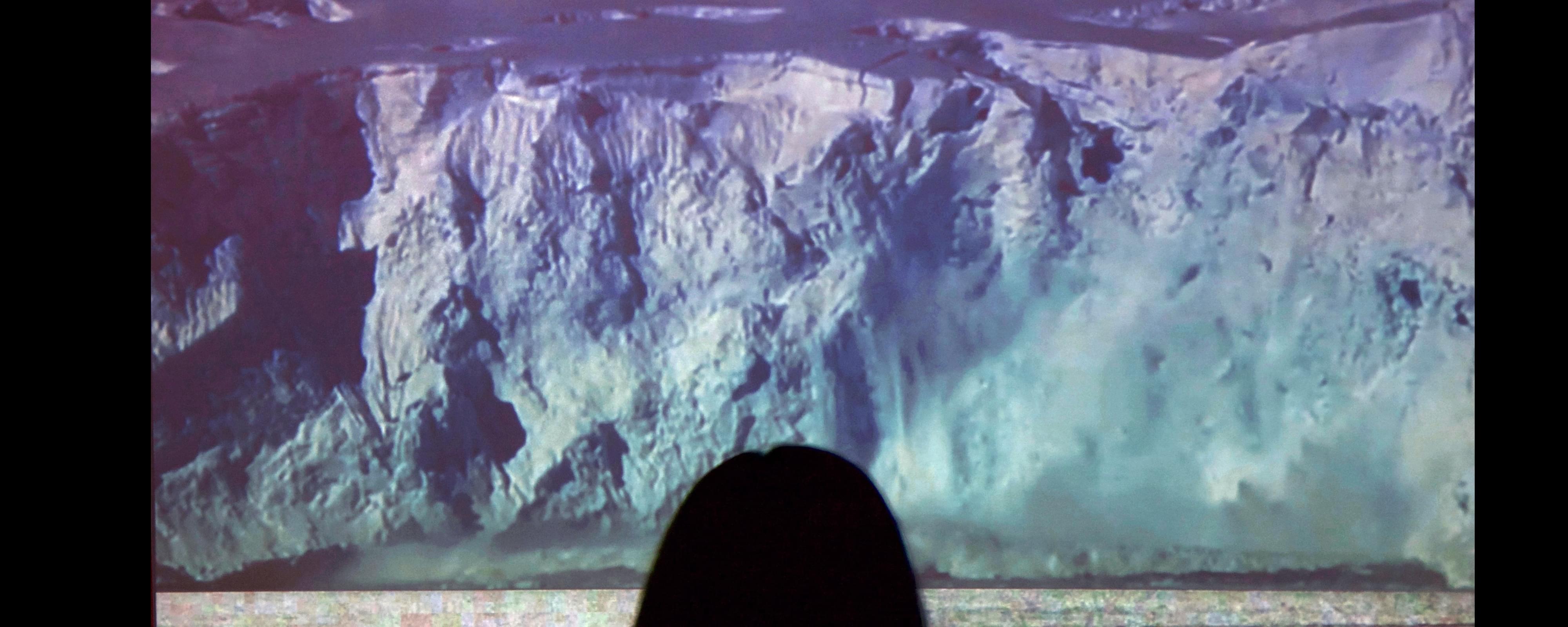 Close-up photograph of the back of a gallery-goer's head as they look at a projected image of a large glacier calving in front of them