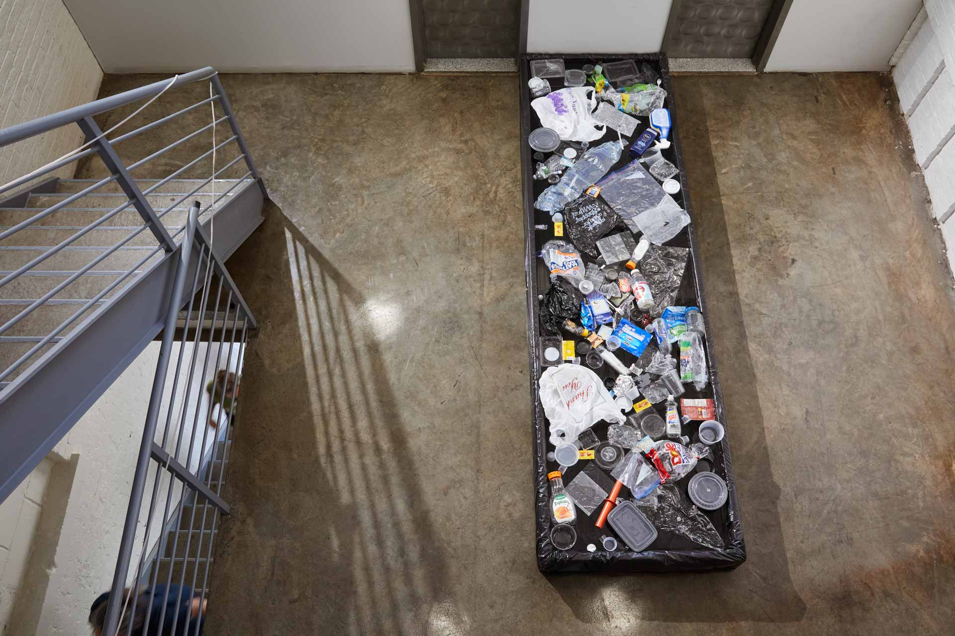 Photograph of top-down view taken from a top a staircase. On the floor below is a ten by three foot small pool, one foot high, filled with water and over a hundred single use plastic items. The pool has a black lining so the water appears black. 