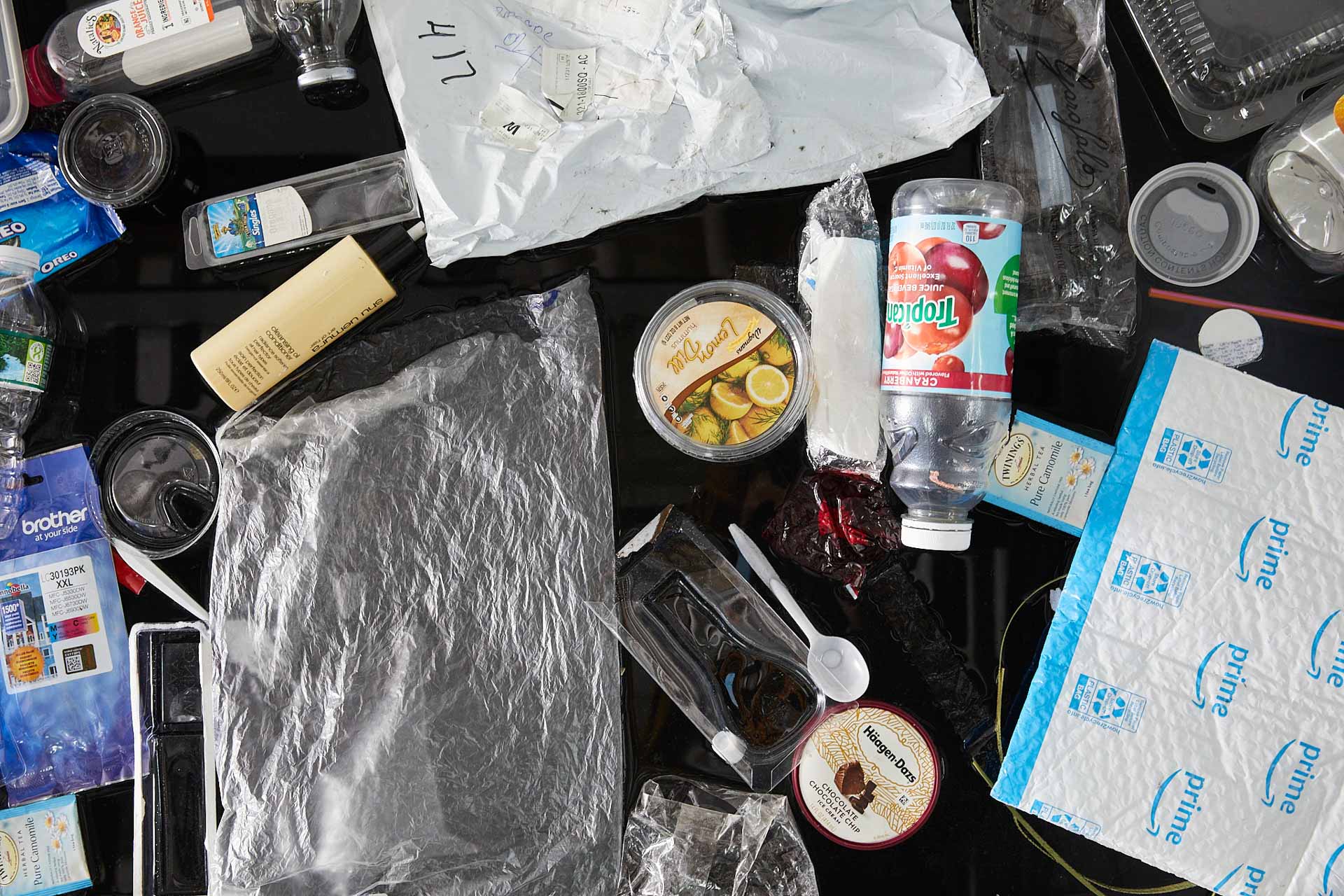 Another tight close up photograph of about 25 single-use plastic items floating on black water. Most prominently featured is a Tropican cranberry juice bottle, Lemon dill hummos container, and a Chocolate Chocolate Chip Haagen Dazs lid.