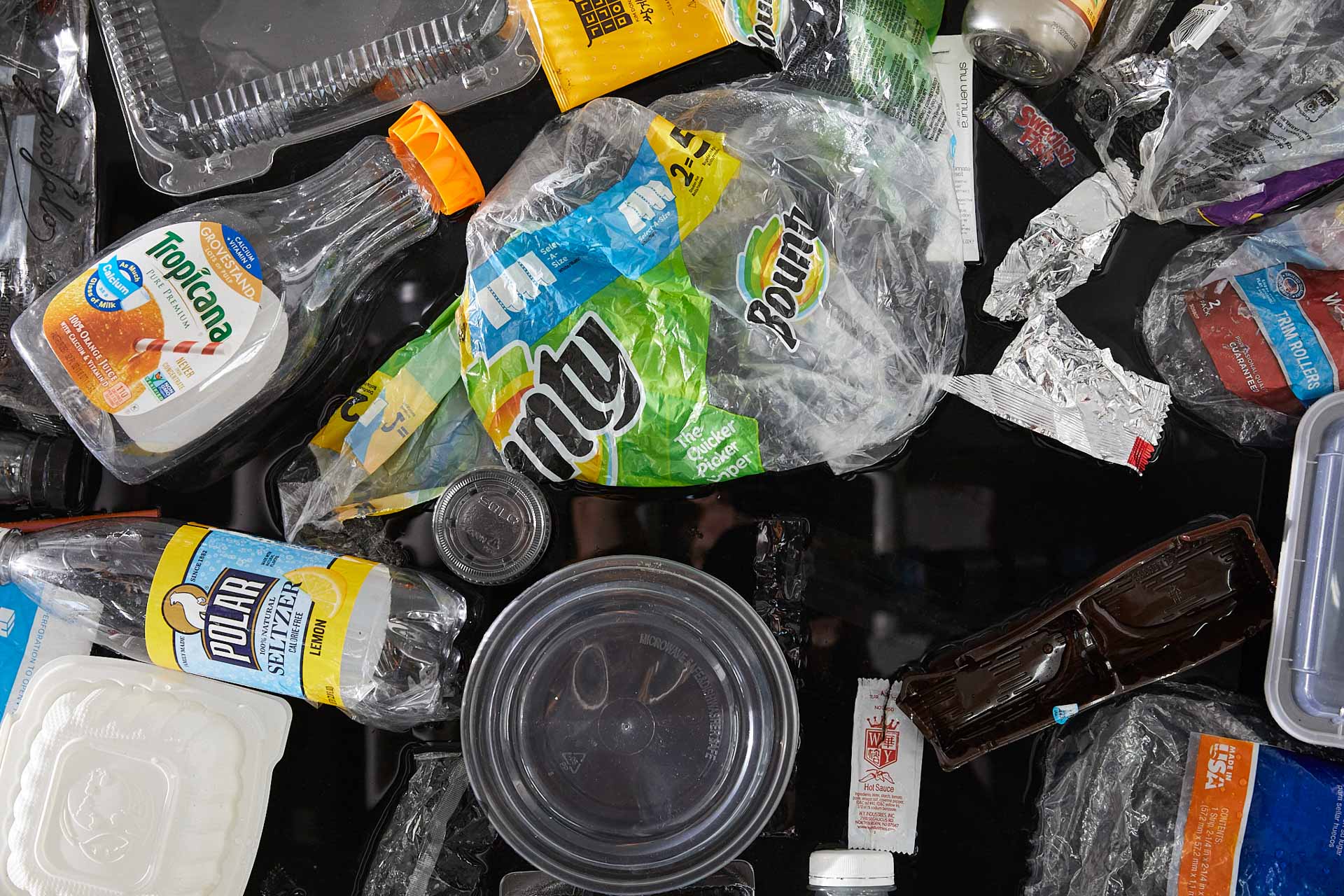 Small thumbnail image of Tight close-up photograph of about 20 single-use plastic items jumbled together. Most prominently featured is a Bounty plastic package wrapping, a Tropican organge juice bottle, and a circular take out container.