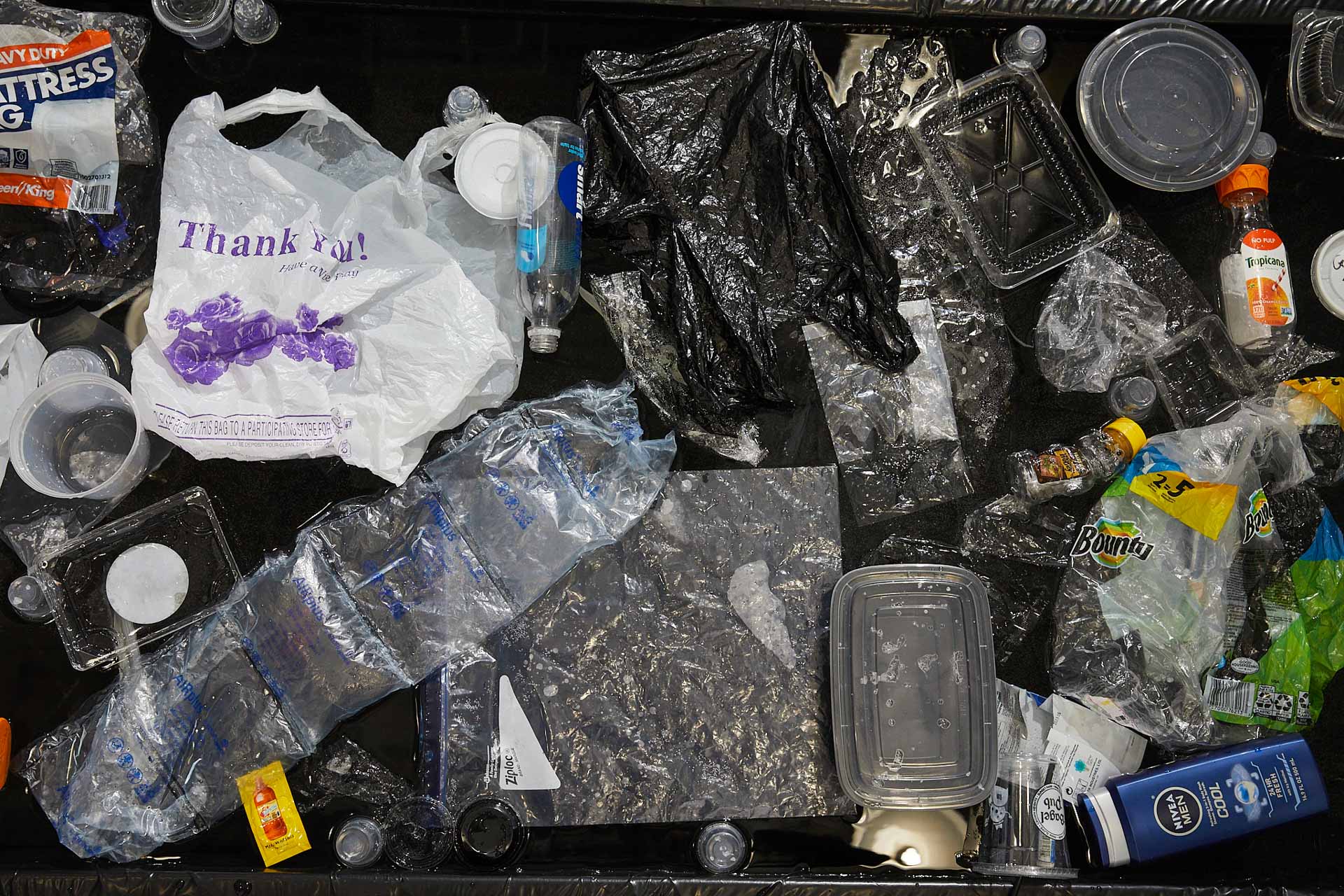 Close-up, top-down photograph of about three dozen single-use plastic items floating on a black body of water. Most prominently featured is a white plastic 'Thank You' shopping bag.