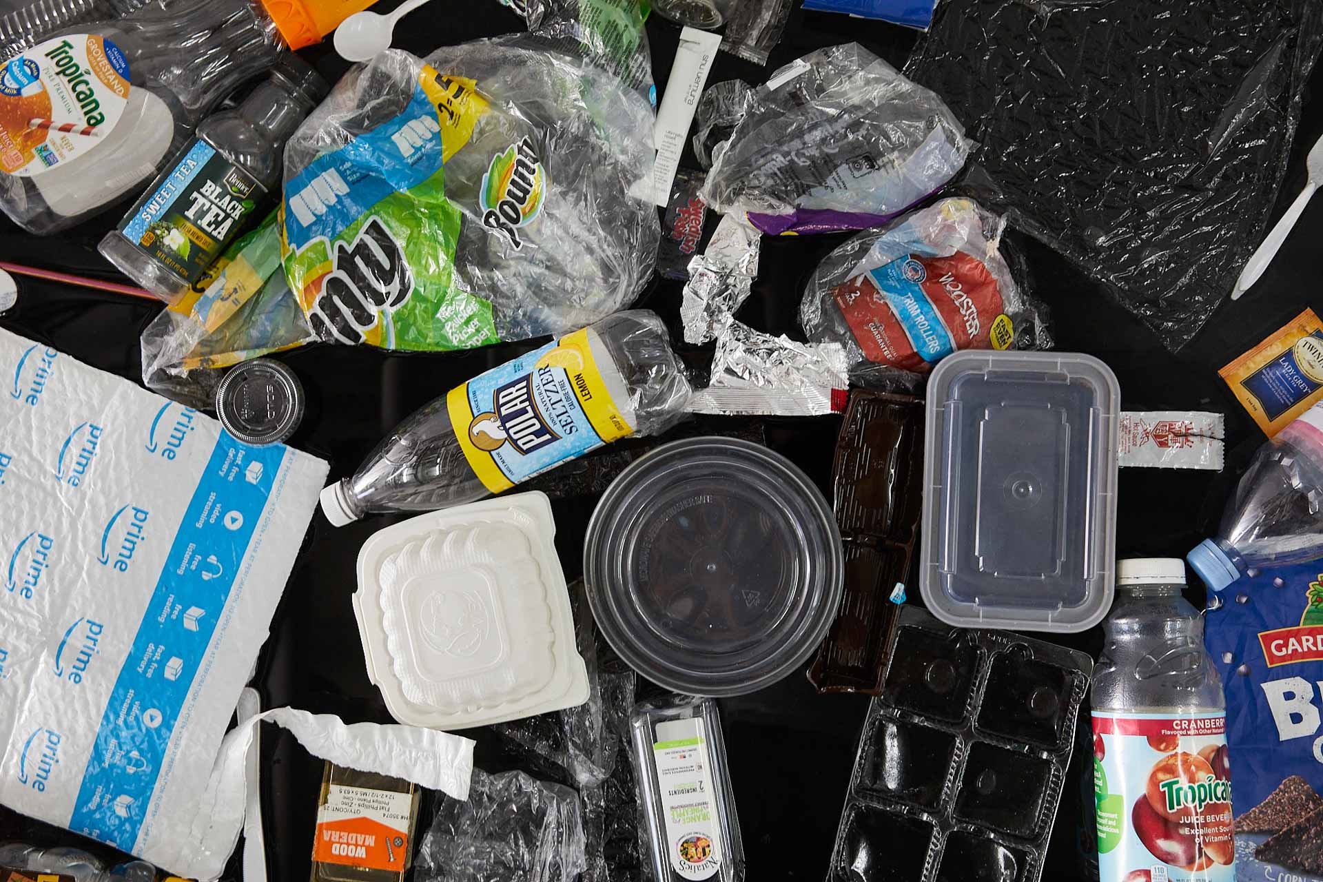 Top-down close-up photo of about 50 pieces of trash floating in a black pool. Most prominently featured is a yellow Polar Seltzer bottle, Bounty plastic wrapping, and an Amazon Prime shipping package.