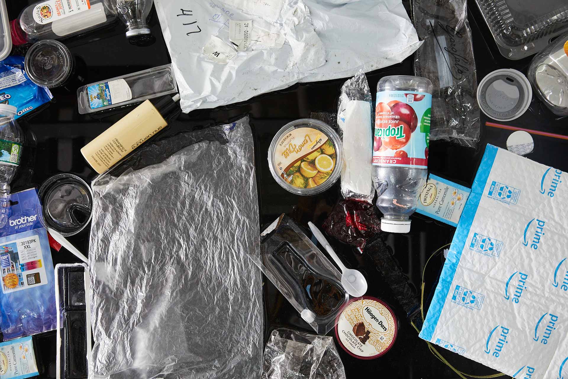 Another tight close-up photograph of about 25 single-use plastic items floating on black water. Most prominently featured is a Tropicana cranberry juice bottle, a lemon dill hummus container, and a Chocolate Chocolate Chip Häagen-Dazs lid.