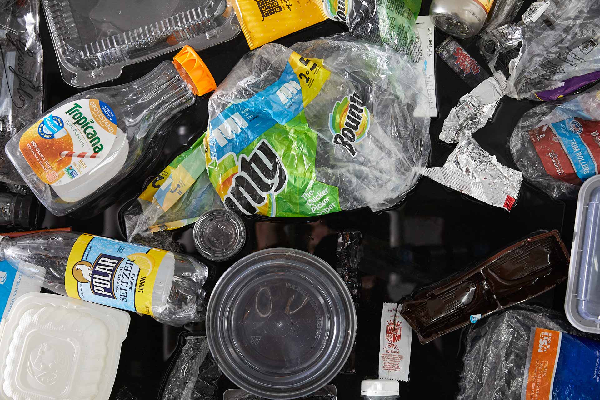 Tight close-up photograph of about 20 single-use plastic items jumbled together. Most prominently featured is a Bounty plastic package wrapping, a Tropicana orange juice bottle, and a circular take-out container.