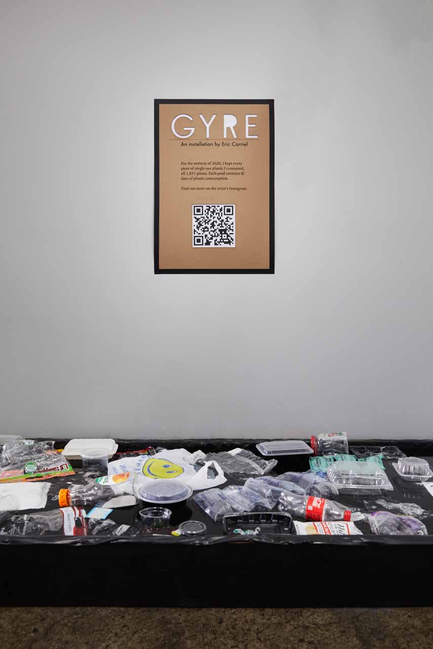 On a gray wall hangs an 18-inch by 24-inch sign that says 'GYRE' followed by some text and a QR code underneath. On the floor, perpendicular to the frame is a one-foot-high black pool with dozens and dozens of single-use plastic items floating on the surface.