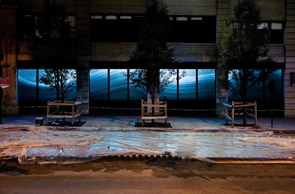 Nighttime photograph showing expansive, 80-foot-long storefront windows with a projection of a geometric rendering of a snowy, bluish, hilly winter landscape that contiguously spans almost the entire width of the storefront. In the foreground, we see a Brooklyn sidewalk fully under construction, with plastic sheeting fully covering the entirety of its surface and a line of &ldquo;CAUTION&rdquo; tape spanning the full length of the frame.