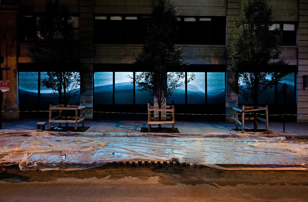 Nighttime photograph showing expansive, 80-foot-long storefront windows with a projection of a geometric rendering of a snowy, bluish, hilly winter landscape in a different geometric configuration than before, which contiguously spans almost the entire width of the storefront. In the foreground, we see a Brooklyn sidewalk fully under construction, with plastic sheeting fully covering the entirety of its surface and a line of &ldquo;CAUTION&rdquo; tape spanning the full length of the frame.