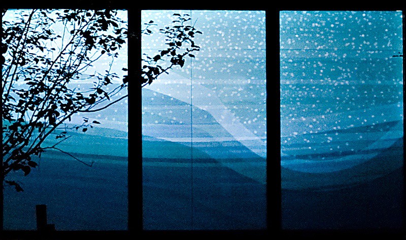 Close-up nighttime photograph of three large storefront windows showing a projection of an undulating geometric rendering of a snowy, bluish, and hilly winter landscape. On the left side of the photo, in front of the projection, are branches emanating from a tree without any leaves.