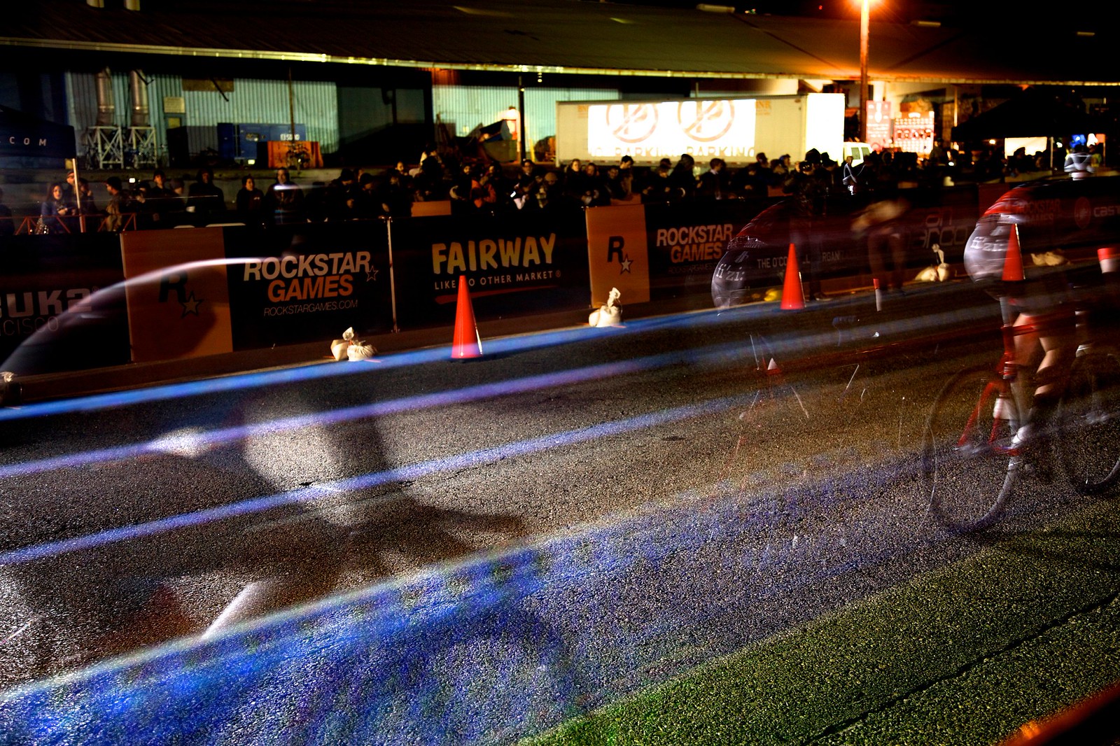 Nighttime, long-exposure photograph showing the &ldquo;light lines&rdquo; of two racing cyclists with a large crowd behind them. Onto the street on which they race is projected a linearly oriented rainbow road of colored lanes.