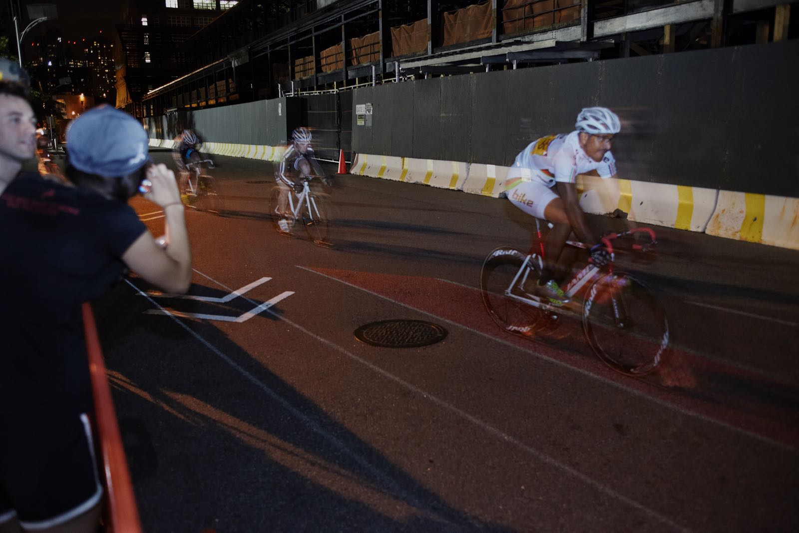 Nighttime photograph of three cyclists, one behind the other, racing on a street course. Underneath the lead cyclist's bike is a thin, illuminated red lane.