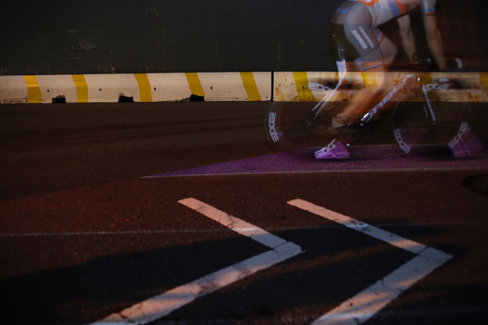 Nighttime, close-up photograph of a street with a thin purple lane projected on it. Riding over the purple lane is a cyclist, whose white cycling shoe is also covered in purple light.