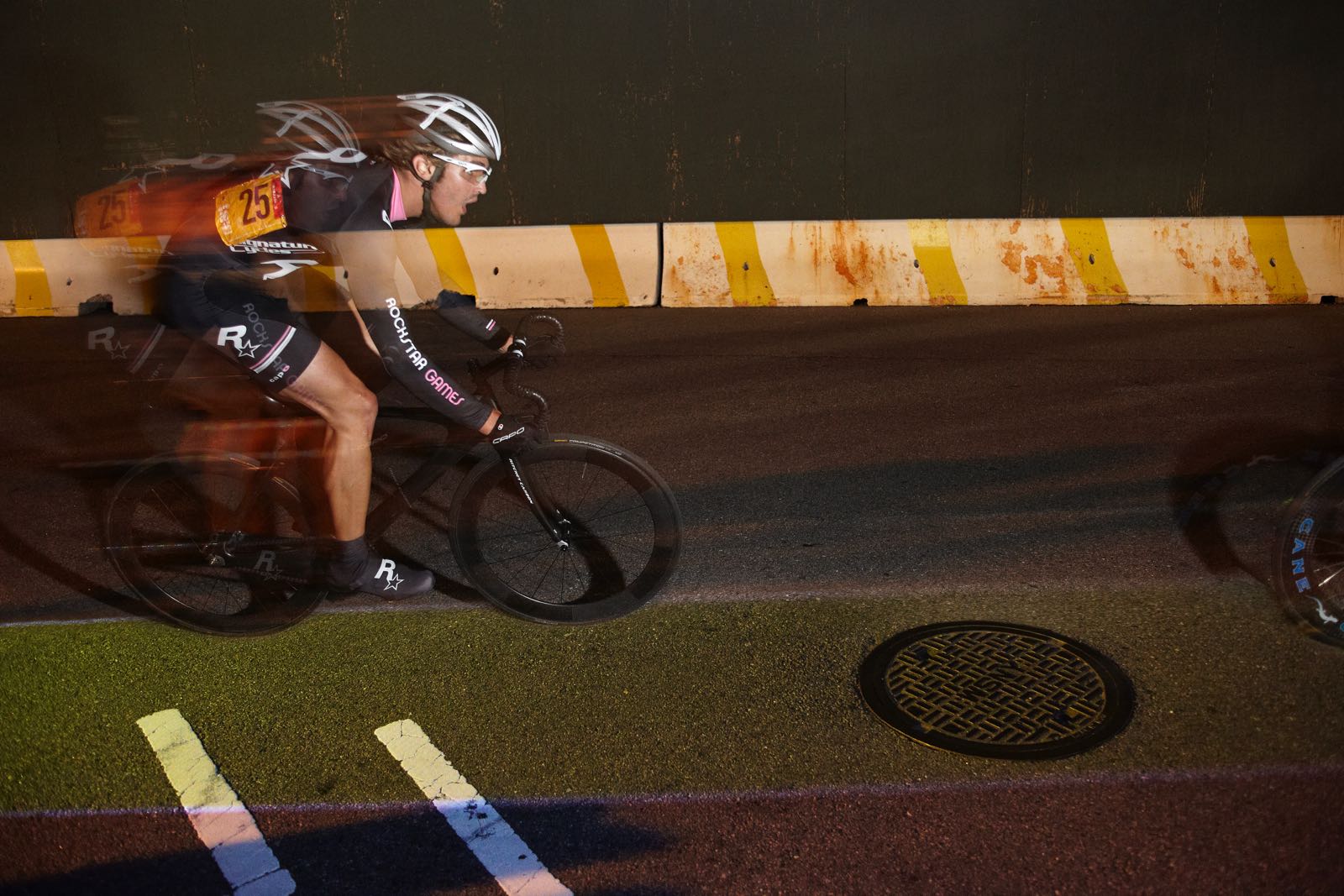Nighttime photograph of one cyclist, seen from the side, racing on a street course. Underneath their bike is a thin, illuminated yellow lane.