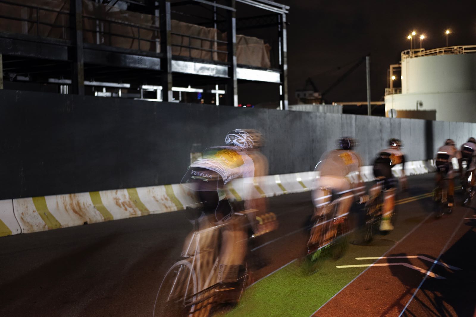 Nighttime, long-exposure photograph showing the 'light lines' of five racing cyclists with a large crowd behind them. Onto the street on which they race is projected a linearly oriented rainbow road of colored lanes
