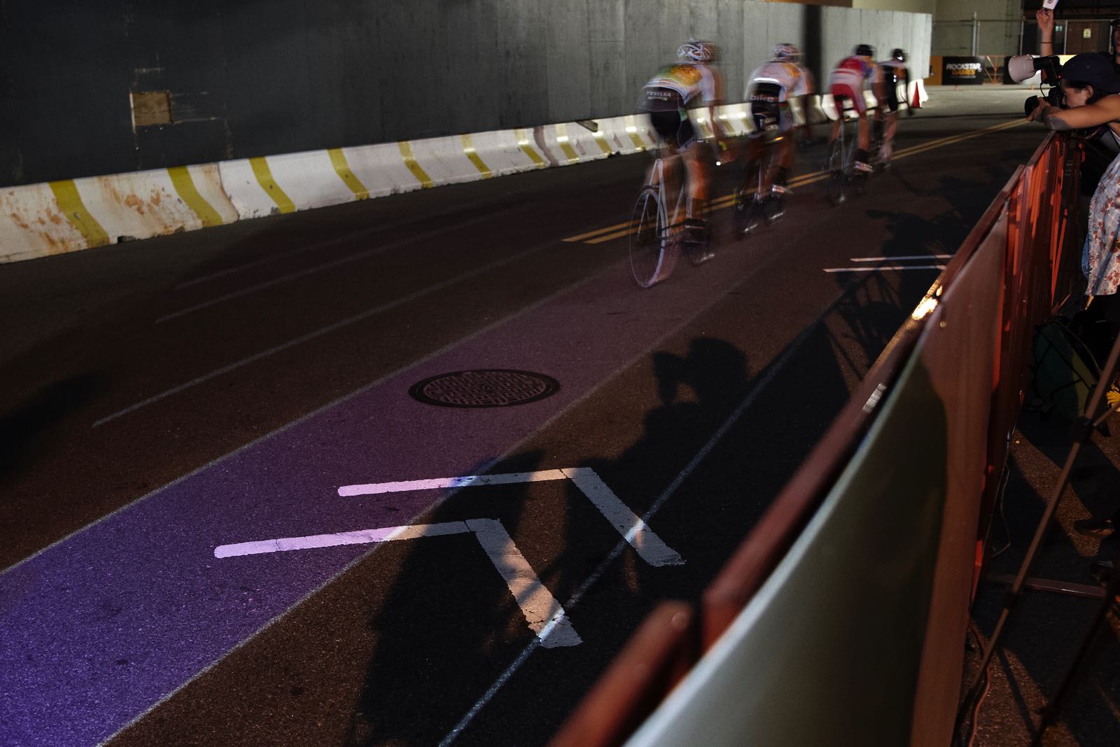 Nighttime photograph of four cyclists, one behind the other, racing on a street course as seen from behind. Underneath the last two cyclists' bikes is a thin, illuminated purple lane.