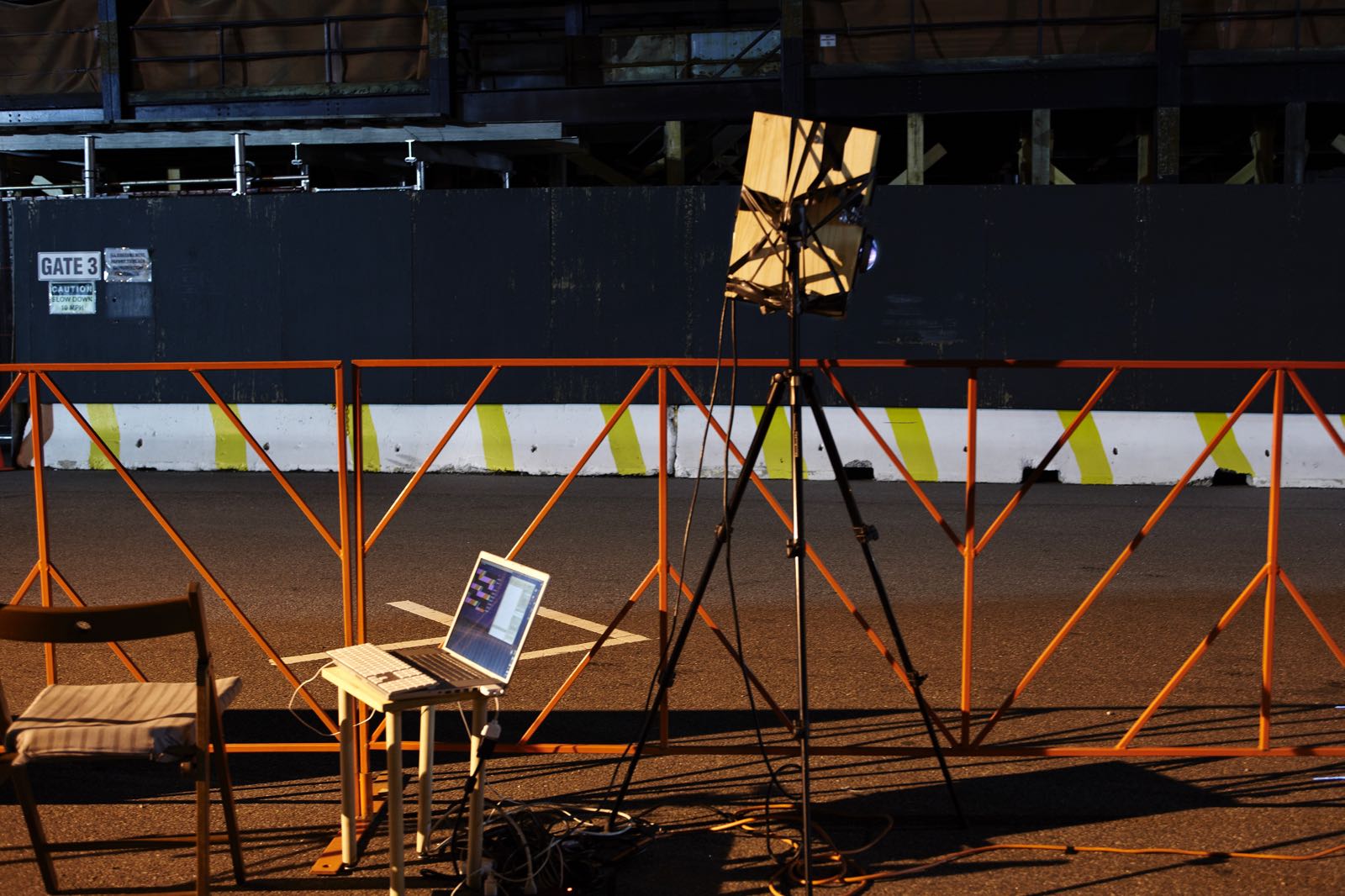 Nighttime photograph showing an orange racing barrier/fence that spans the full length of the frame. In front is a chair with an open laptop on a stand connected to a projector on a high tripod. In the background is an empty race course. There aren't any people in the photograph.