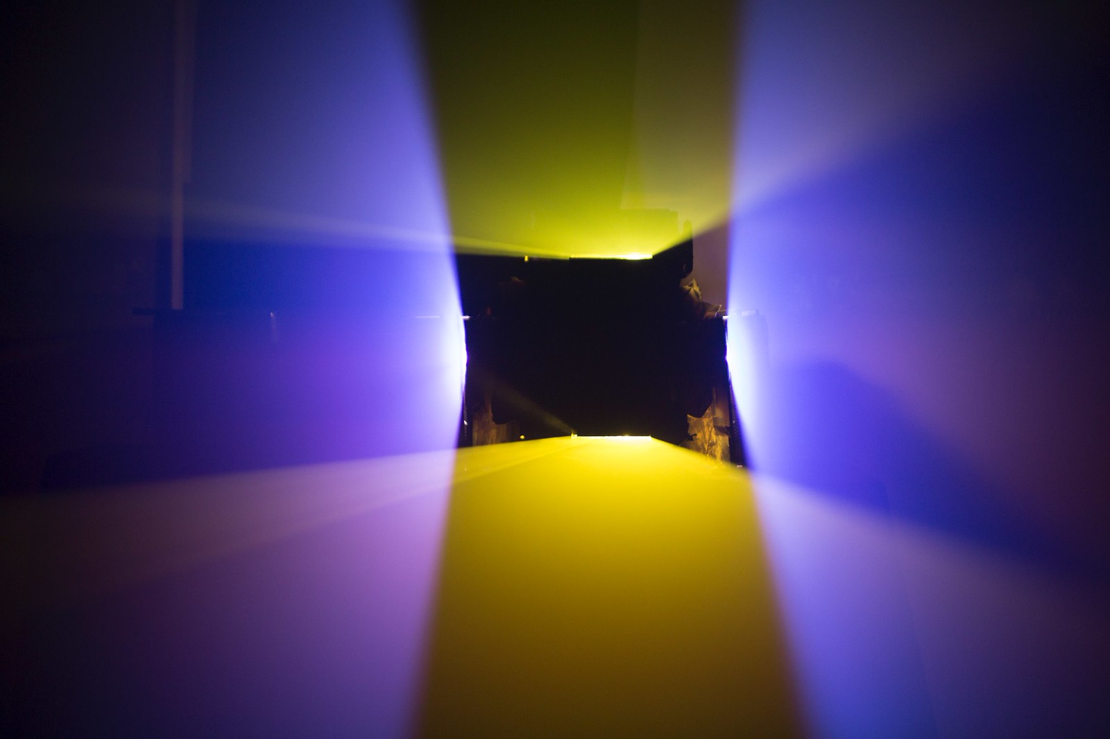 Photograph of a dark room lit by two vertically oriented yellow planes of light and two horizontally oriented purple planes of light. The planes intersect each other to form a black, hollow square shape (where there isn&rsquo;t any light) in the middle of the frame.