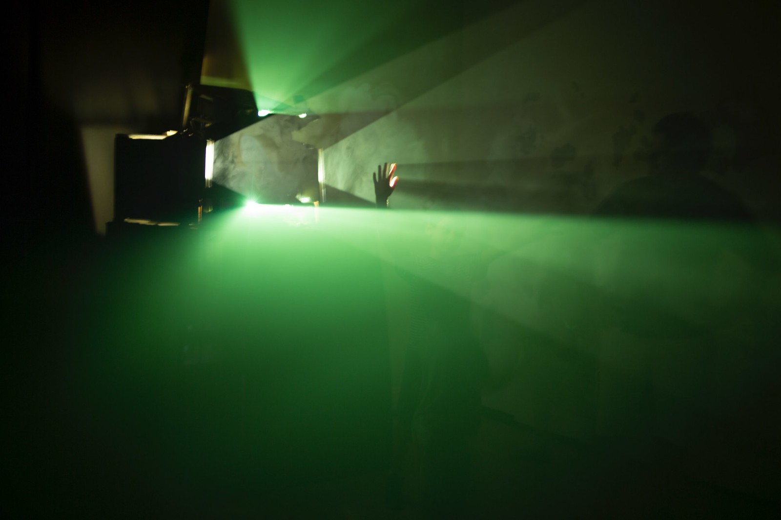 Photograph of a dark room with a strong light source (projector) on the left side of the frame. Emanating from the projector, in a fog-filled room, are very prominent and sharply divided planes of pure green light, perfectly parallel to the floor/ceiling and about 18 inches apart. In the gap stretches a child&rsquo;s hand, which creates its own sharply defined shadows in the opposite direction of the light source.