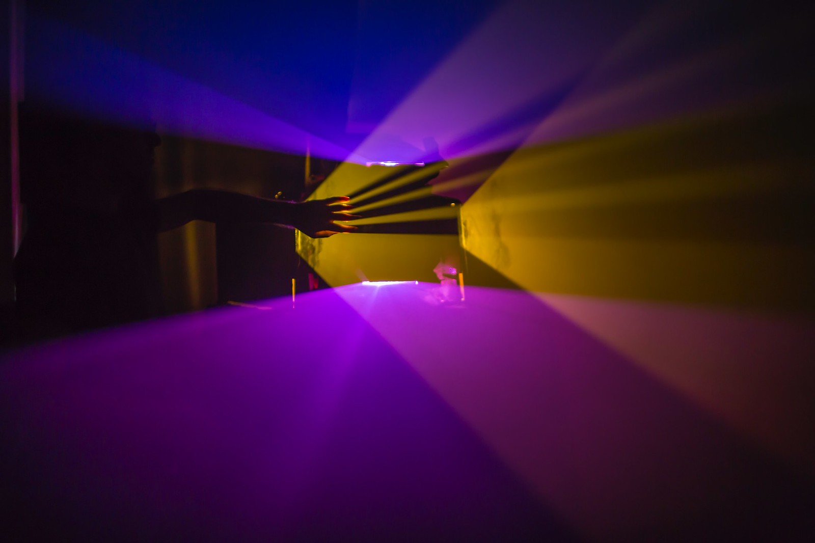Photograph of a dark room with a strong light source (projector) on the left side of the frame. Emanating from the projector, in a fog-filled room, are very prominent and sharply divided planes of purple and orange light that form V-shapes as they expand outward. In the middle of the frame, a hand reaches up from the shadows and creates its own sharply defined shadows in the opposite direction of the light source.