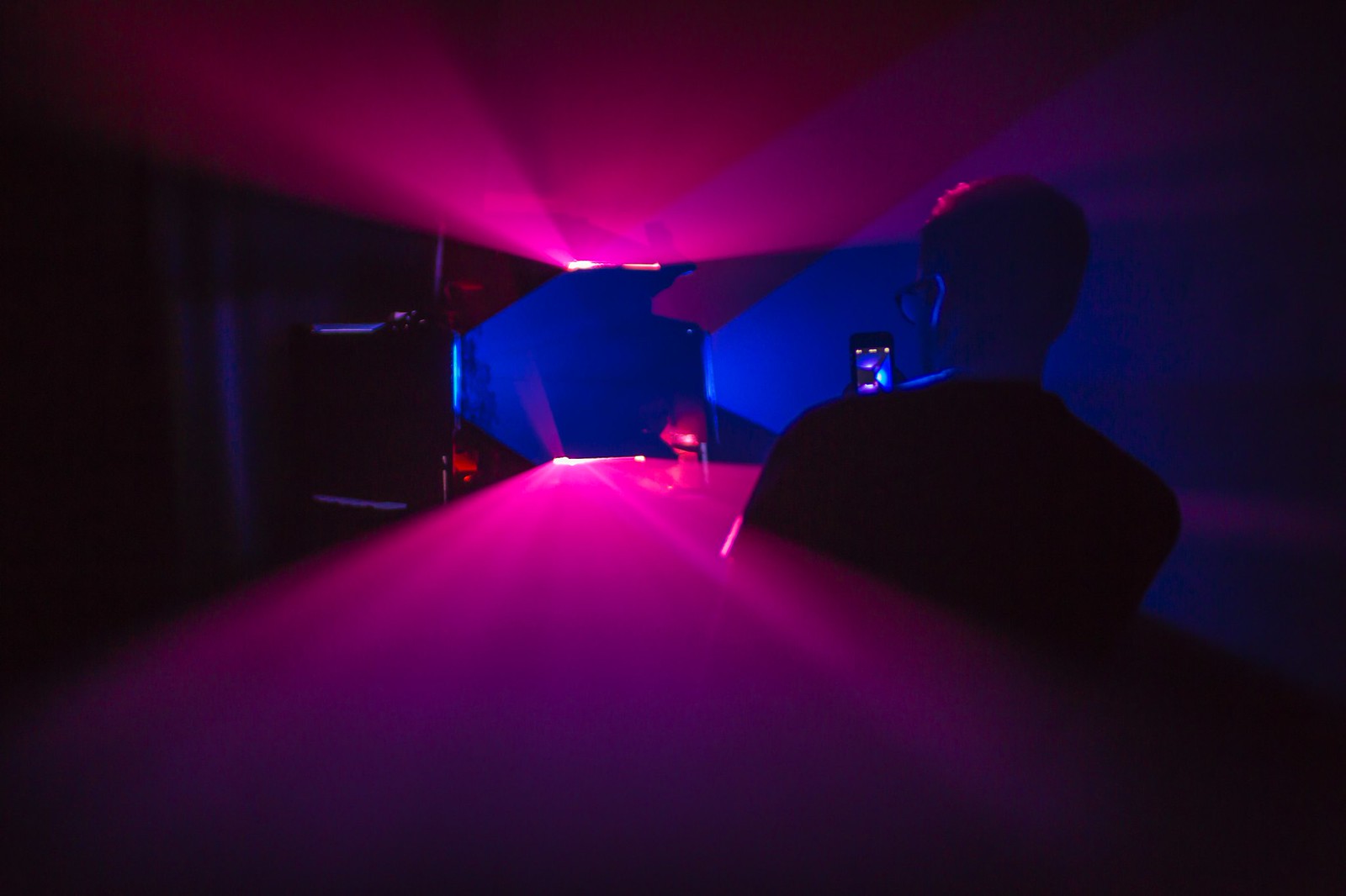 Photograph of a dark room lit by two vertically oriented lavender planes of light and two horizontally oriented yellow planes of light. The planes intersect each other to form a black, hollow square shape (where there isn&rsquo;t any light) in the middle of the frame.