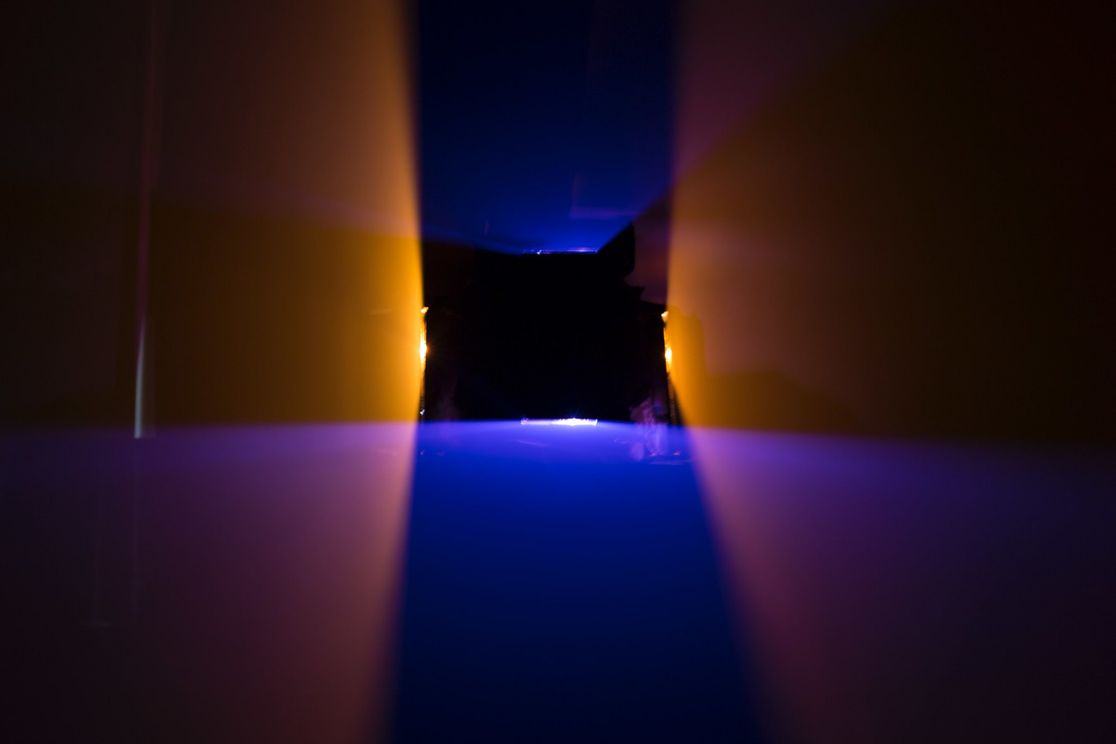 Photograph of a dark room lit by two vertically oriented orange planes of light and two horizontally oriented blue planes of light. The planes intersect each other to form a black, hollow square shape (where there isn&rsquo;t any light) in the middle of the frame.