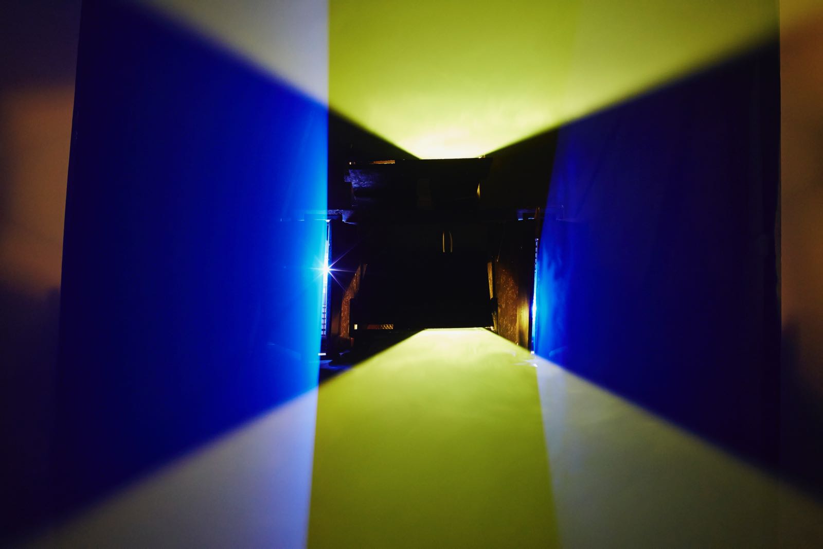 Photograph of a dark, fog-filled room, in the middle of which hover two rectilinearly bound, horizontal planes of yellow light. The planes are roughly 18 inches apart, and in the background is a small crowd of gallery-goers.