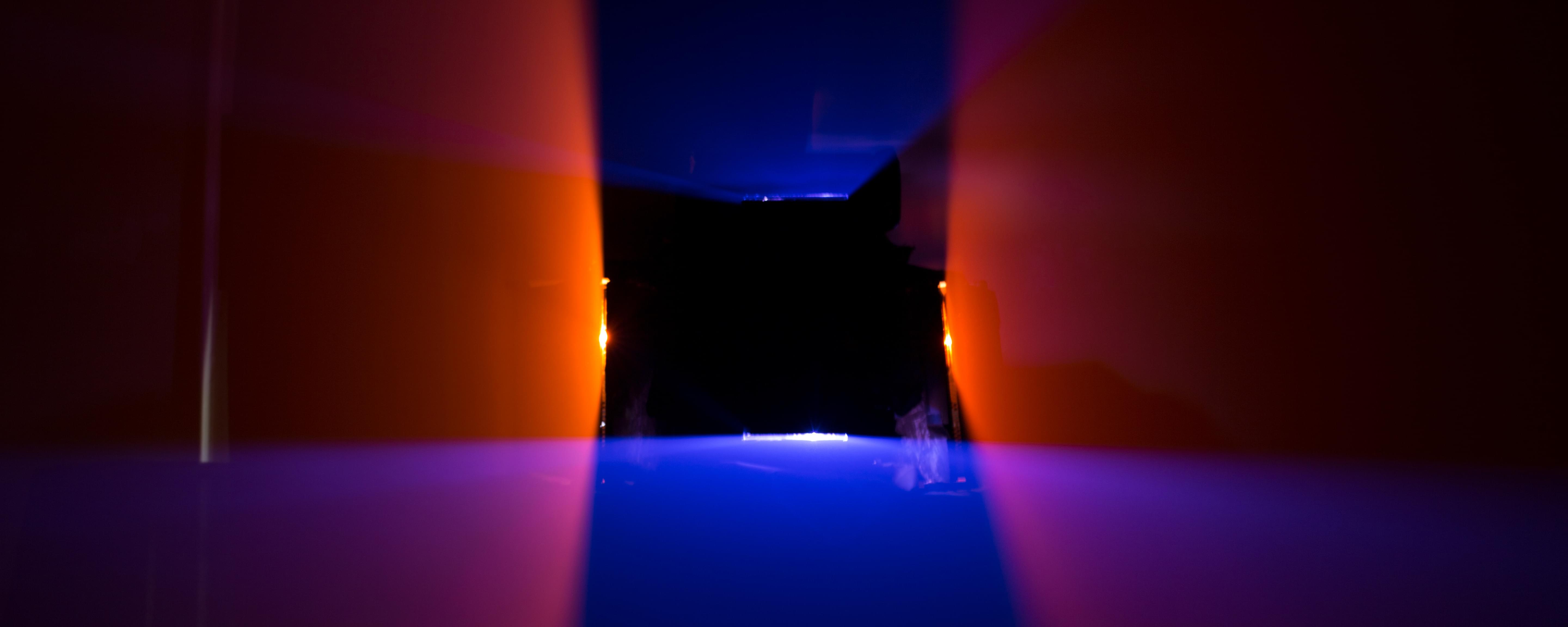 Very strange photograph of a rectangular black hole in the middle, about the size of a cafeteria lunch tray. Emanating from the top and bottom of the rectangular black hole are blue rhomboid planes of light that get wider as they approach the viewer. Emanating from the left and right side of the rectangular black hold are red-orange rhomboid planes of light that also get wider as they approach the viewer.