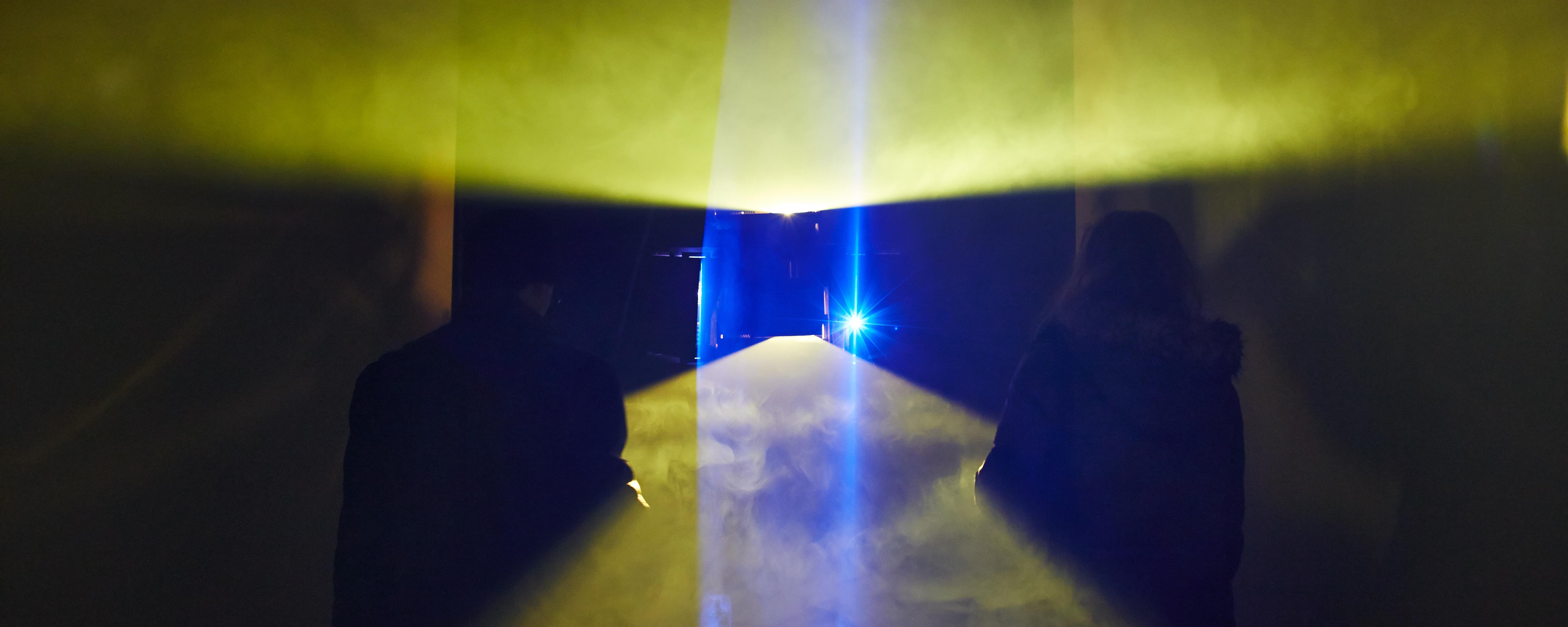 Photograph; planes of yellow light fan out from the top and bottom of the frame; a rectangular black hole in the middle; blue planes of light fan out from the sides. The planes of light reveal fog in the air. Two silhouettes of people with their backs to the camera are probably wondering what the fuck is going on.