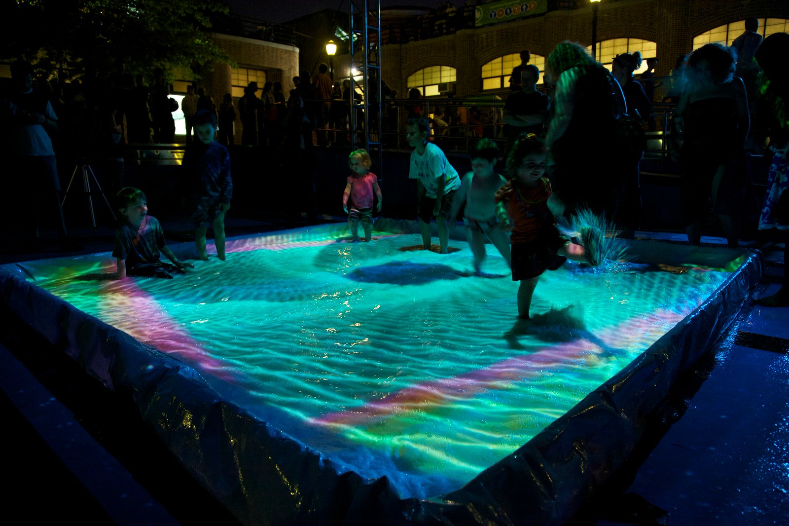 Nighttime photograph of a group of children playing in a small pool, the surface of which is being projected onto with a teal green geometric pattern