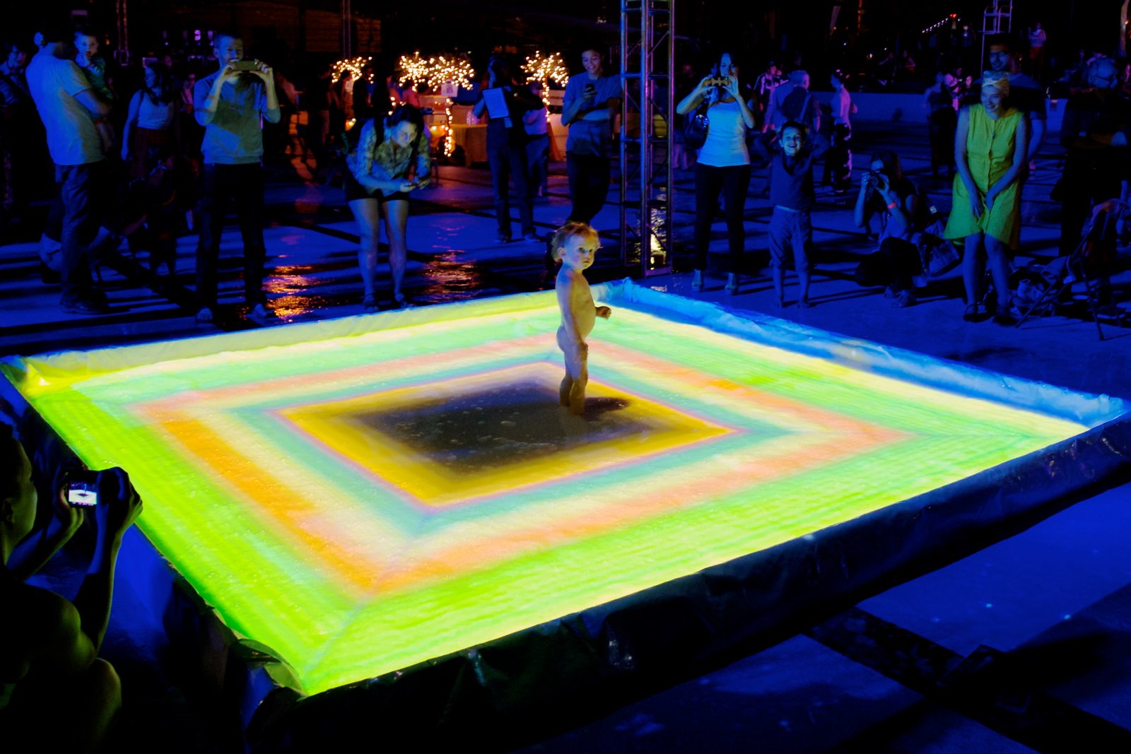 Nighttime photograph of a naked toddler standing in the middle of a small pool, the surface of which is being projected onto with a green, orange, and yellow pattern of concentric rectangles
