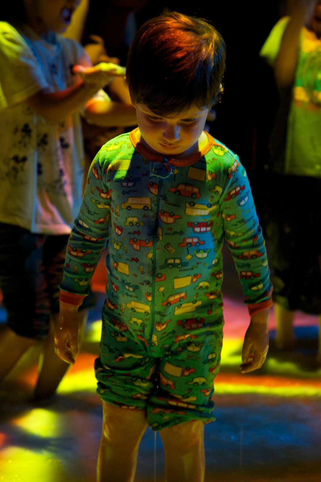 Nighttime photograph of a small child in pajamas looking down is illuminated by an orange light emanating from the colorful surface of the pool he is standing in