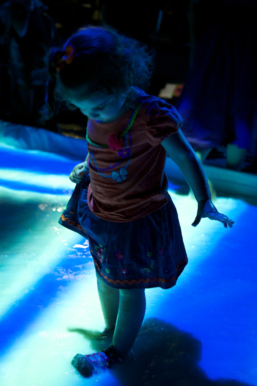 Nighttime photograph of a small girl looking down at her feet which are immersed in a pool of white liquid onto which a blue and pink geometric pattern is projected