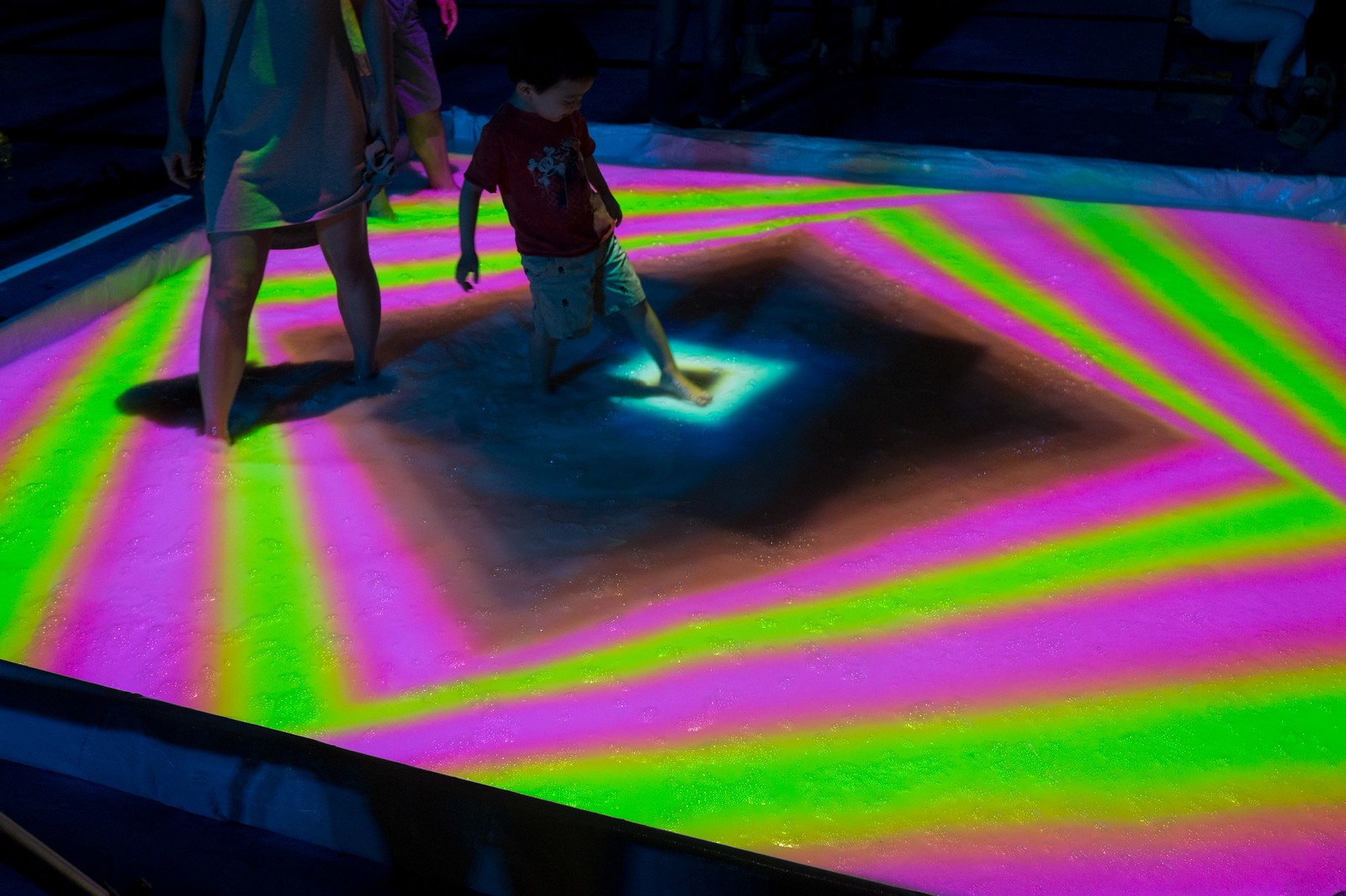 Nighttime photograph of a child in a small pool, the surface of which is being projected onto with a fluorescent pink and green concentric rectilinear pattern with a black diamond at the center, which is where the child is putting their foot