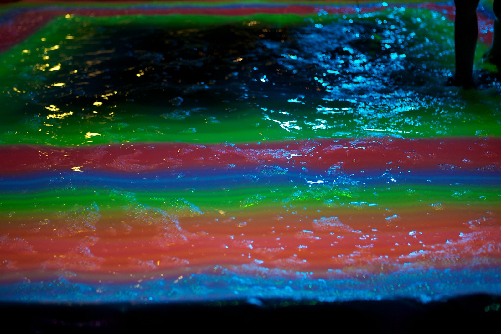 Nighttime close-up photograph of rippling water in a small pool, the surface of which is being projected onto with a geometric rainbow of stacked green, red, blue, and orange rectangles