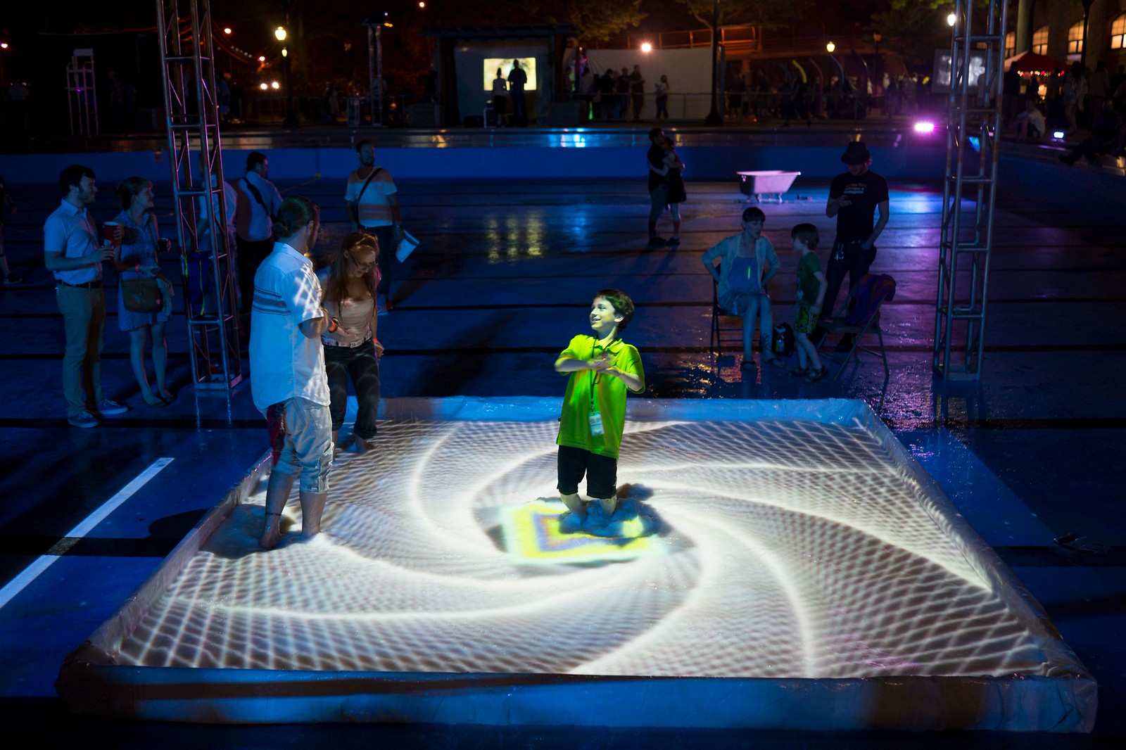 Nighttime photograph of a small pool inside which a small child and an adult play. Projected on the surface is a white spiral of concentric geometric shapes