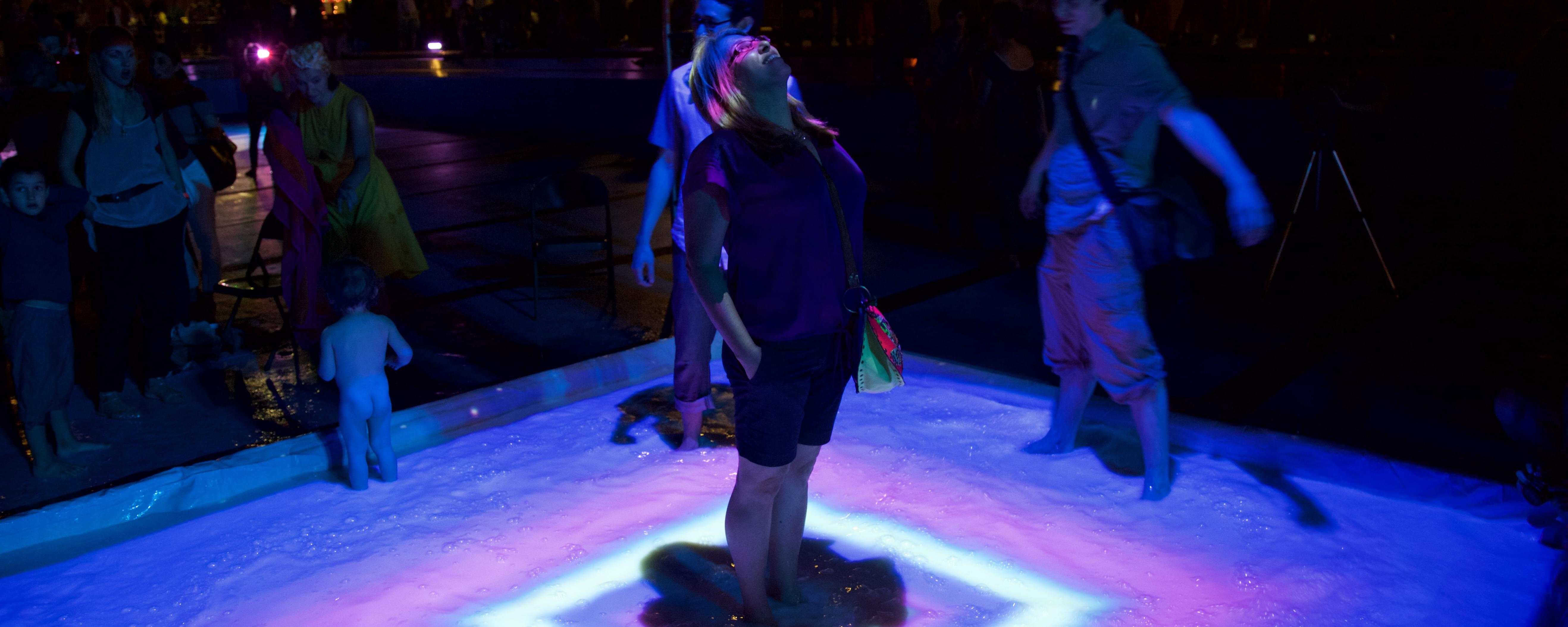 Nighttime photograph of a woman standing in the middle of a 8 inch high pool. Projected from above is a crazy multi-colored lightshow that covers her eyes in a ray of purple light while the liquid she's standing in (milk, btw) catches the purple, blue, and violet rays that fall around her.