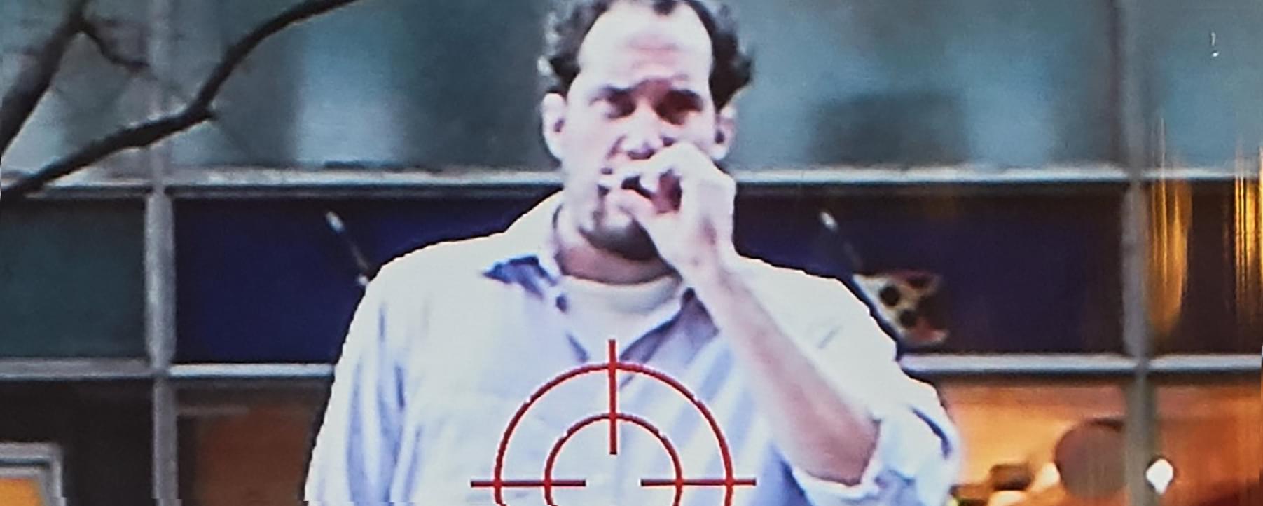 Close up grainy photograph of a man smoking a cigarette in a white button down shirt, with a cross-hairs target symbol shown on top his shirt