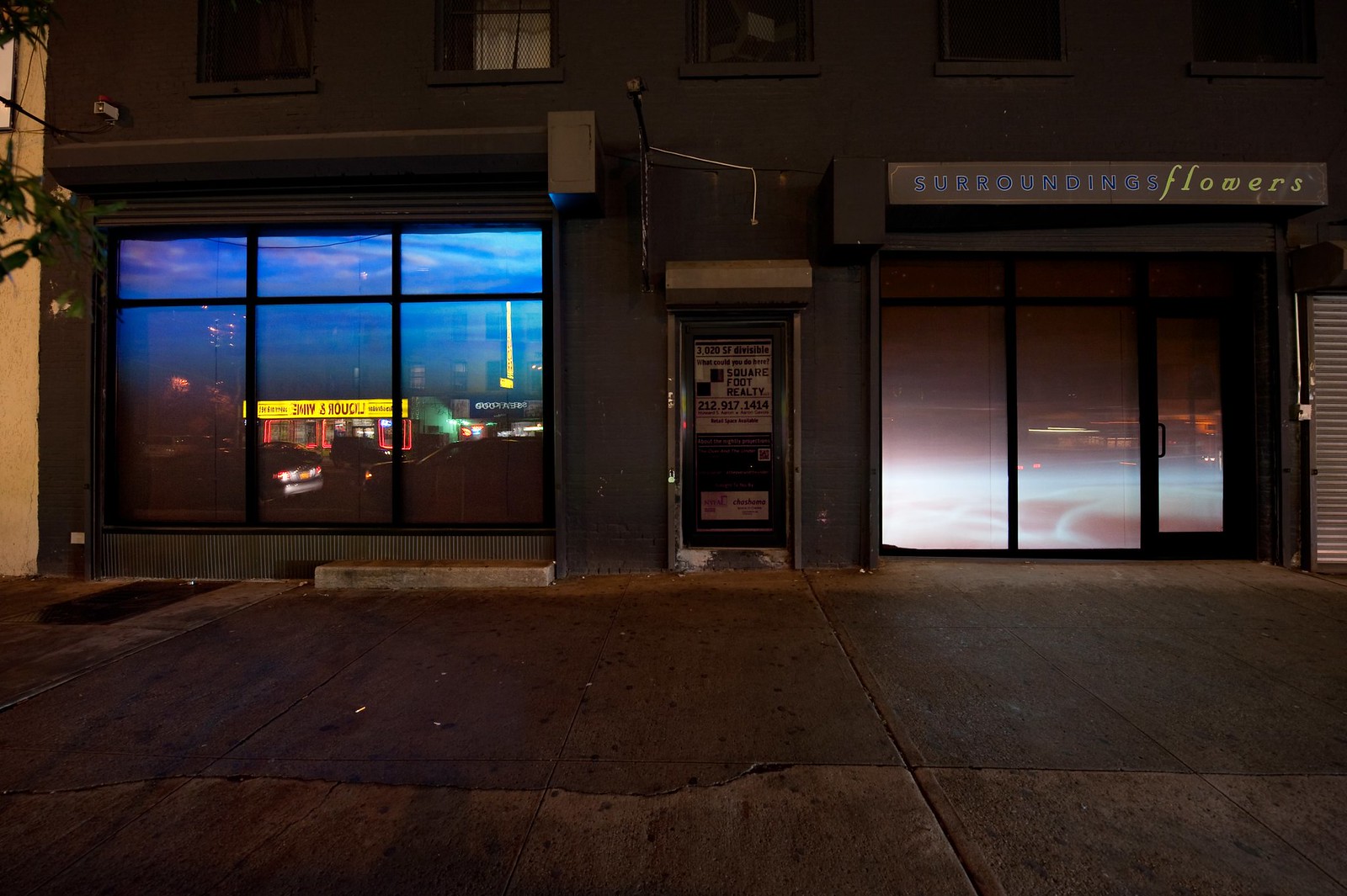 Nighttime photograph showing a wide, urban sidewalk in the foreground and a wide storefront that occupies the full width of the frame in the background. The storefront consists of two large windows on either side separated by a door in the middle. Projected into the leftmost window is a (different) image showing water from beneath the surface and projected in the rightmost window is a pink-hue image of a sky with fluffy clouds effectively resting at sidewalk level.