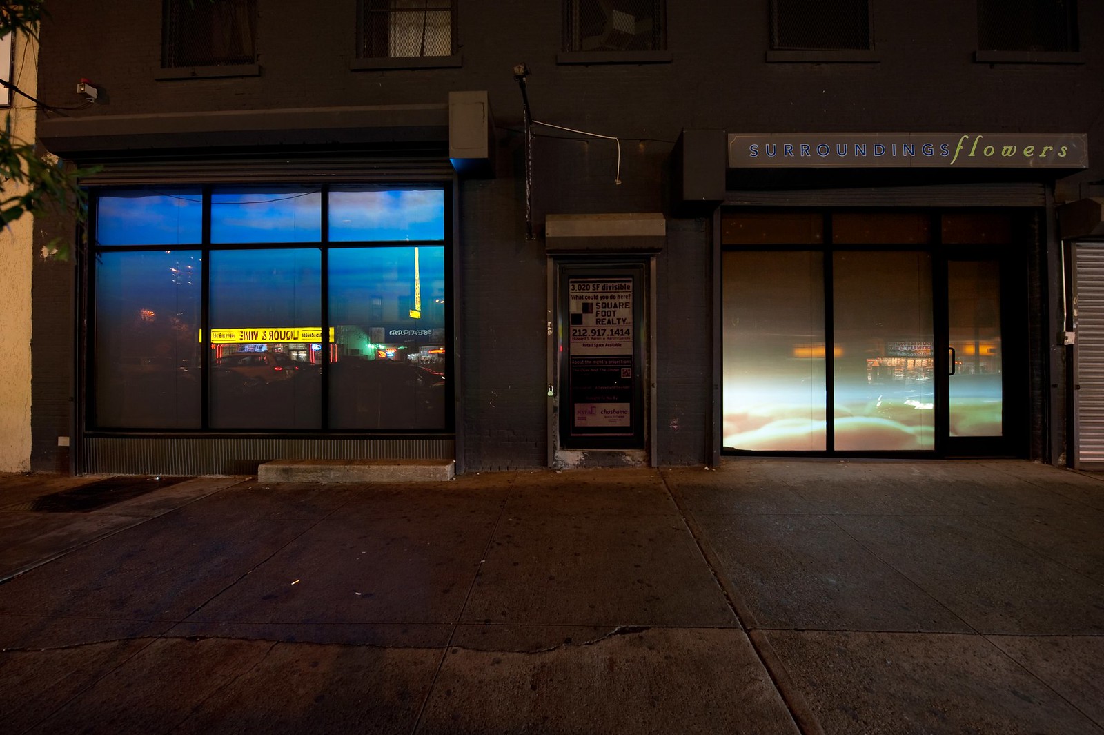 Nighttime photograph showing a wide, urban sidewalk in the foreground and a wide storefront that occupies the full width of the frame in the background. The storefront consists of two large windows on either side separated by a door in the middle. Projected into the leftmost window is a (different) image showing water from beneath the surface and projected in the rightmost window is a golden yellow-hue image of a sky with fluffy clouds effectively resting at sidewalk level.