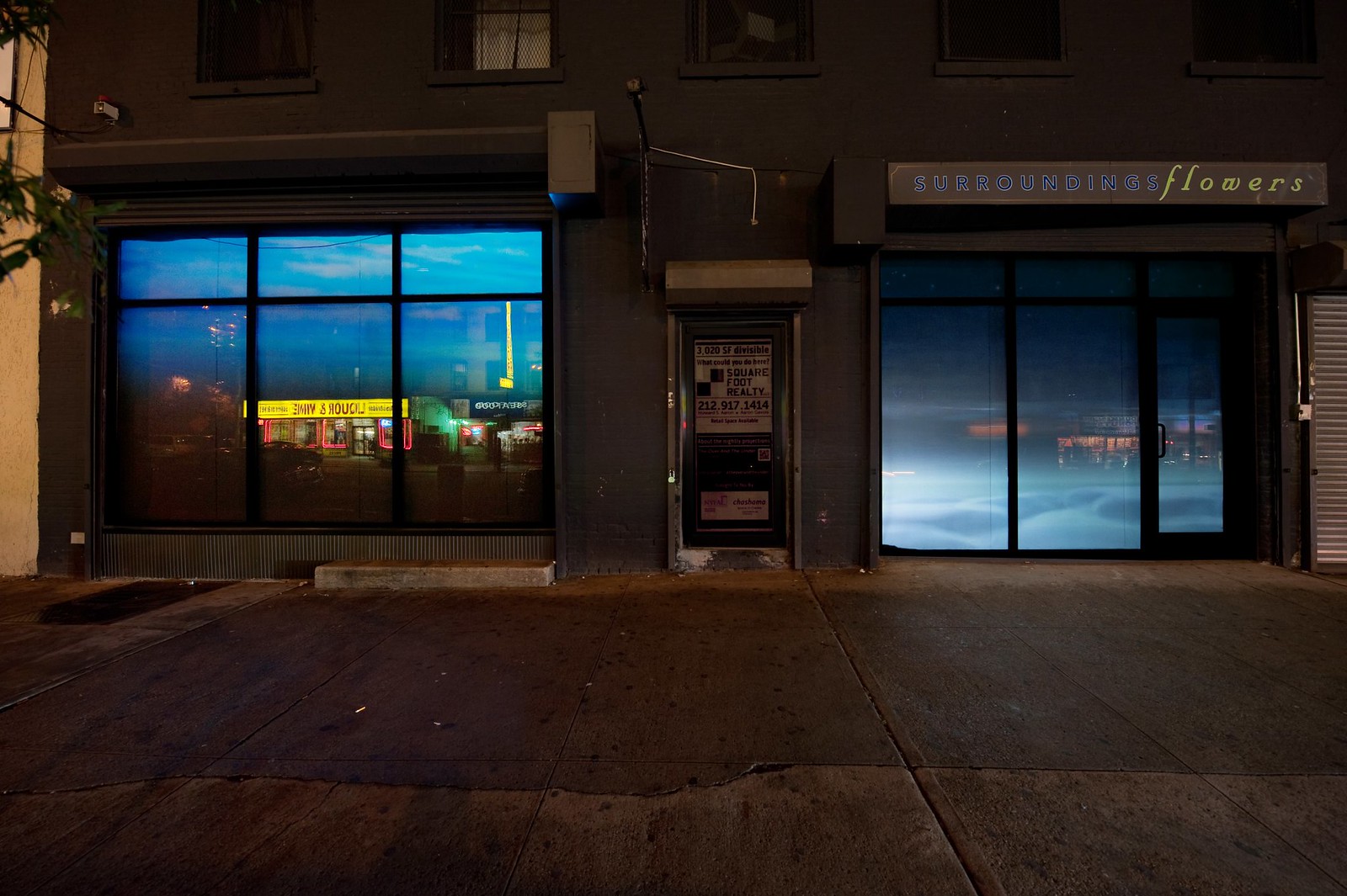 Nighttime photograph showing a wide, urban sidewalk in the foreground and a wide storefront that occupies the full width of the frame in the background. The storefront consists of two large windows on either side separated by a door in the middle. Projected into the leftmost window is a (different) image showing water from beneath the surface and projected in the rightmost window is a blue-hue image of a sky with fluffy clouds effectively resting at sidewalk level.