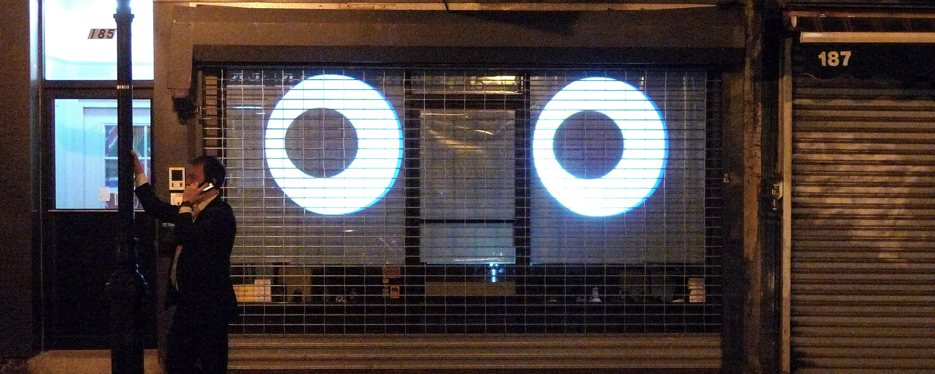 Nighttime photograph of an urban dweller on a cellphone, leaning against a lamppost on the far right side of the frame. Occuping the rest of the frame are two giant cartoon-like eyes, that are projected into the gated storefront behind him. The eyes are eyeing him; he has his back to them, seemingly oblivious to their presence.