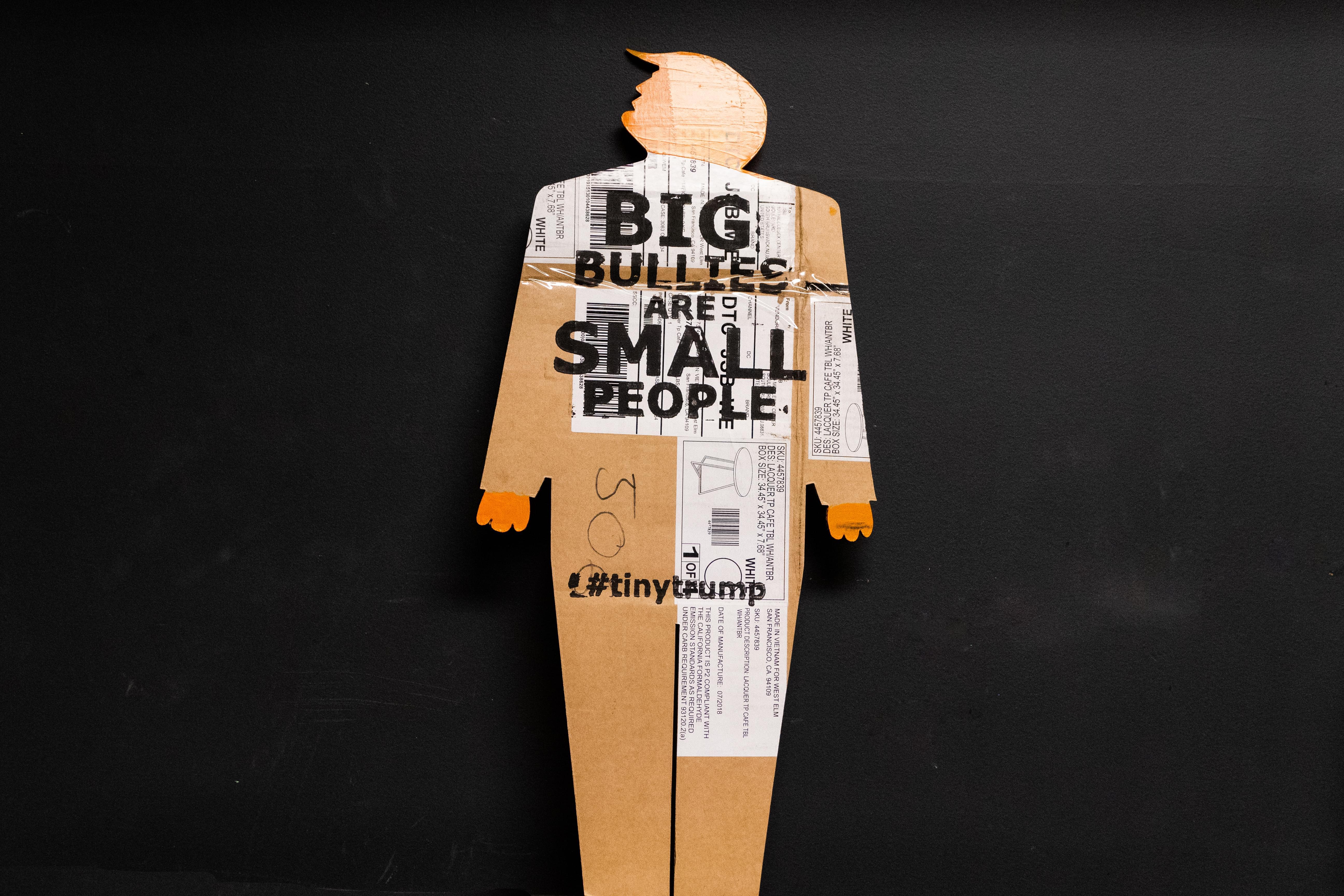Two-foot-tall caricature cardboard cut out of trump with clown like hands and swooshy hair. Head and hands are painted bright orange and the slogan Big Bullies Are Small People is stamped in black ink across the chest. The cardboard is clearly used--it has shipping labels on it. Gray photo studio background.